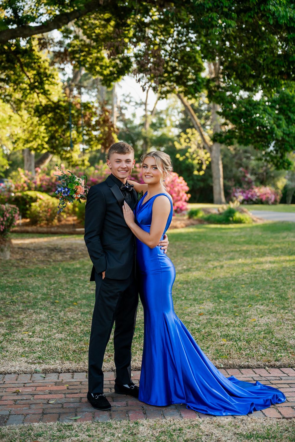 A young couple in formal prom attire, man in black tuxedo and woman in royal blue gown, pose in a lush garden setting.