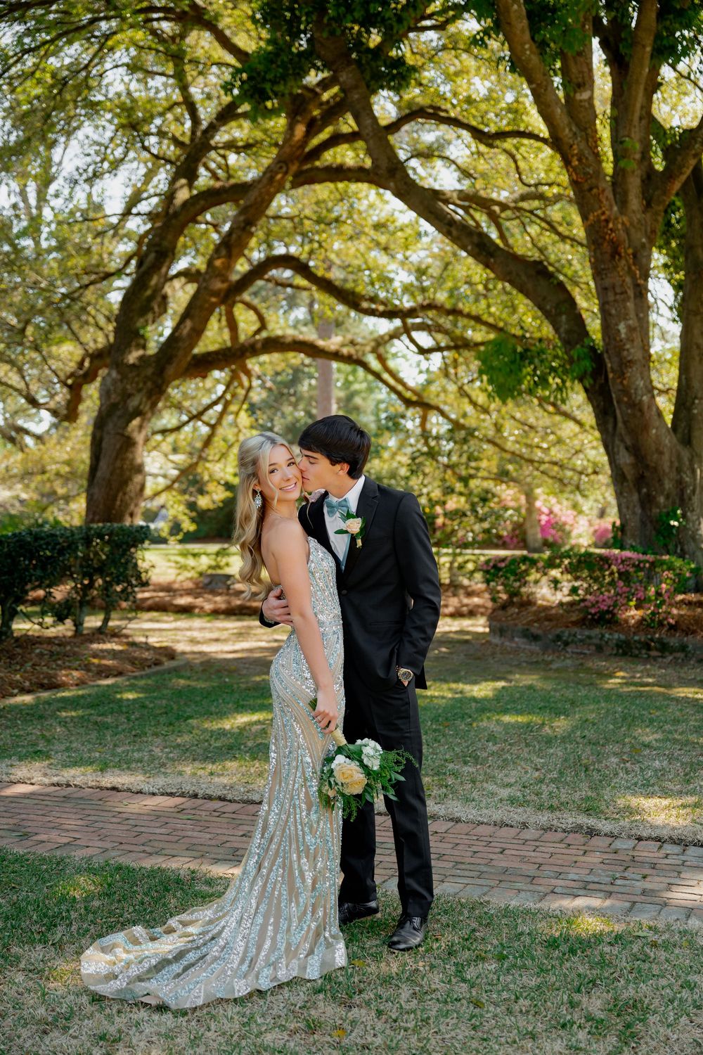 A couple in formal prom attire poses outdoors under large oak trees, the young man kissing her cheek as she holds a bouquet.
