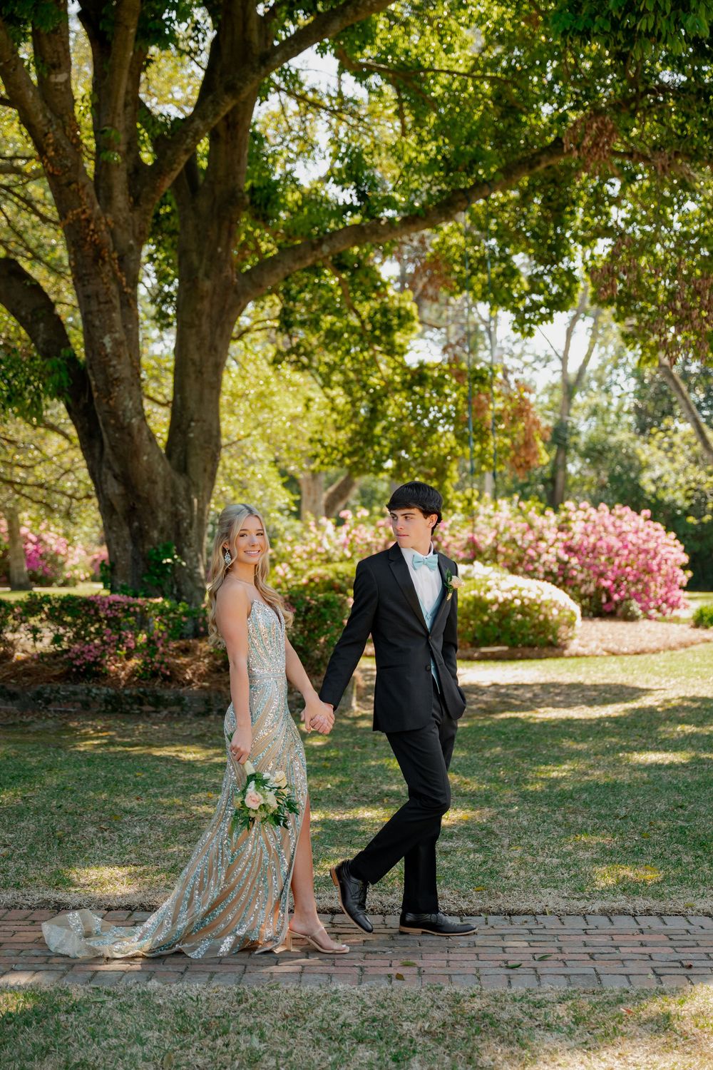 Bride and groom walk hand-in-hand on brick path in lush garden, she wears gold gown, he wears black tuxedo.