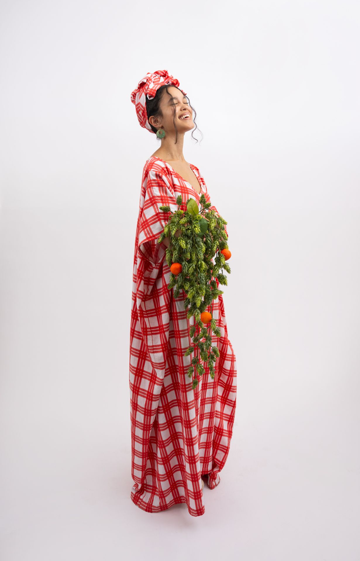 Woman in red plaid kaftan and head wrap holds fresh green herbs with orange tomatoes, smiling joyfully.