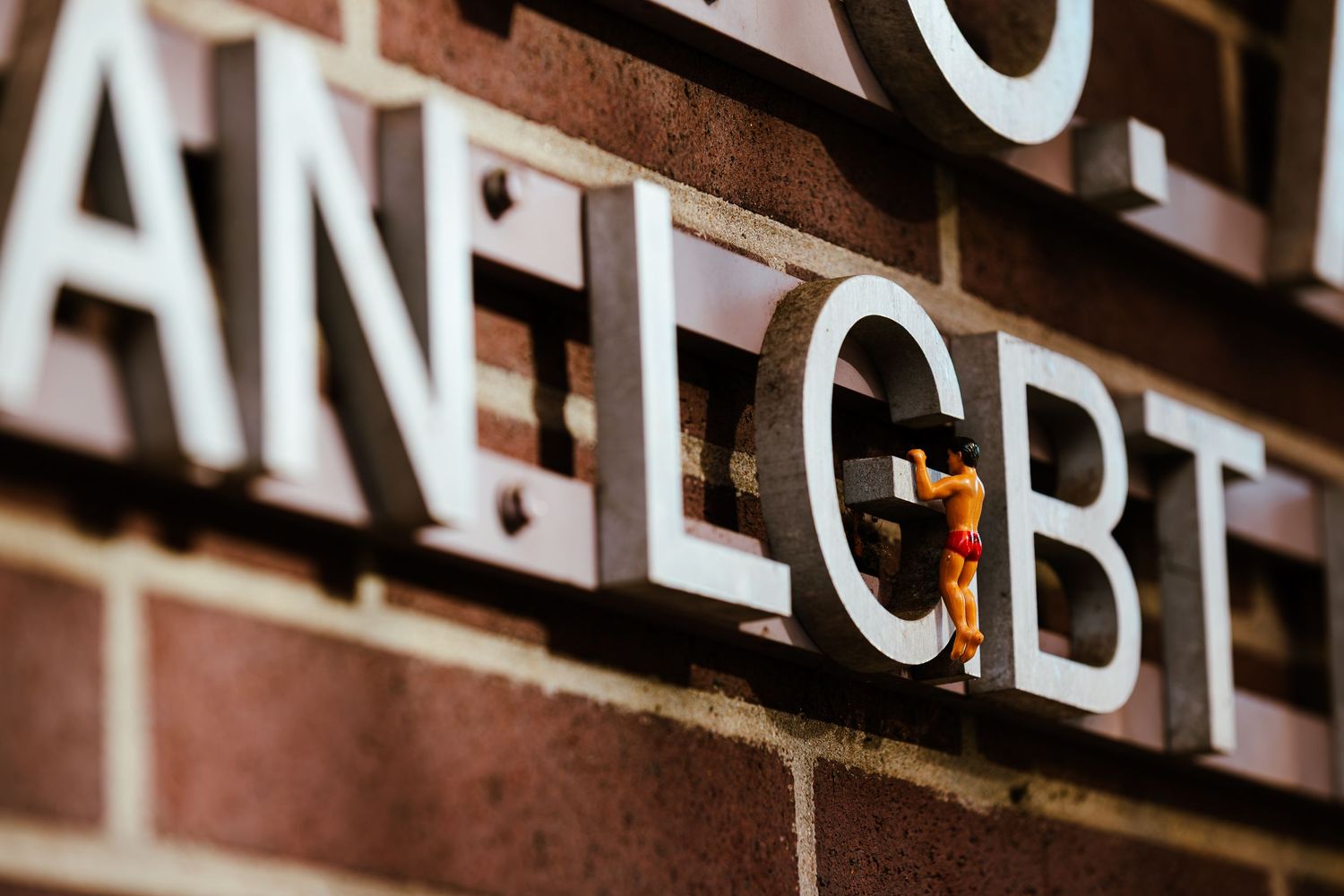 Miniature figurine standing against a brick wall with large LGBT sign letters in close-up perspective.