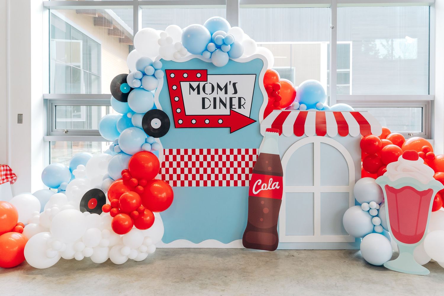 Retro diner-themed party backdrop with balloon arch in red, white and blue featuring a milkshake sign and striped awning.