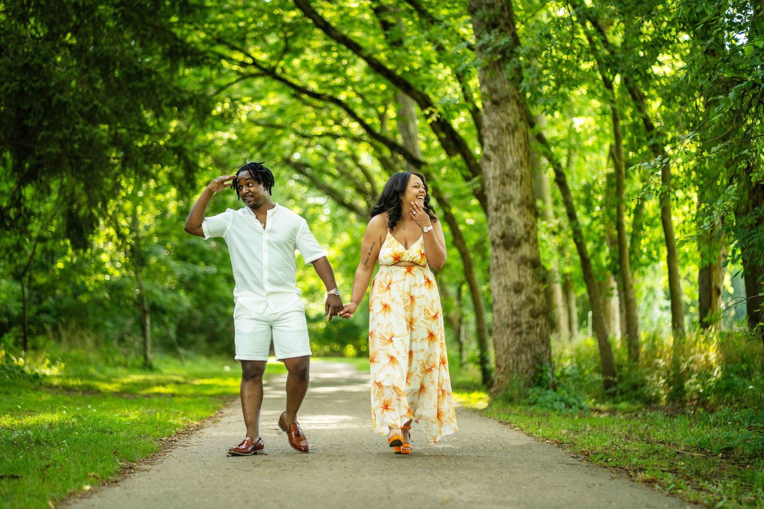 A couple walks hand in hand down a tree-lined path on a sunny summer day, surrounded by lush greenery.