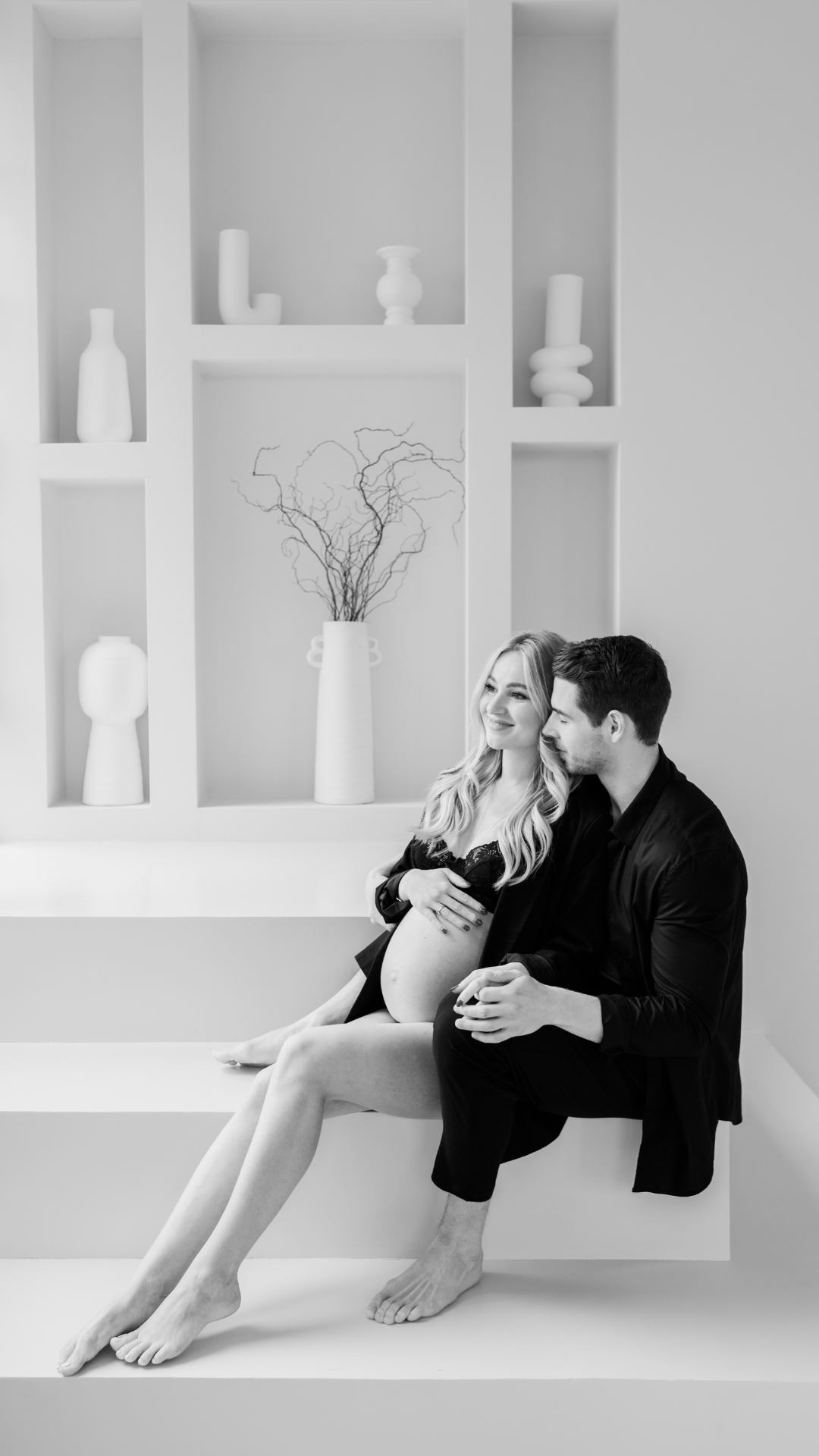 A romantic black and white photograph of a couple embracing in a minimalist white room with built-in shelves.