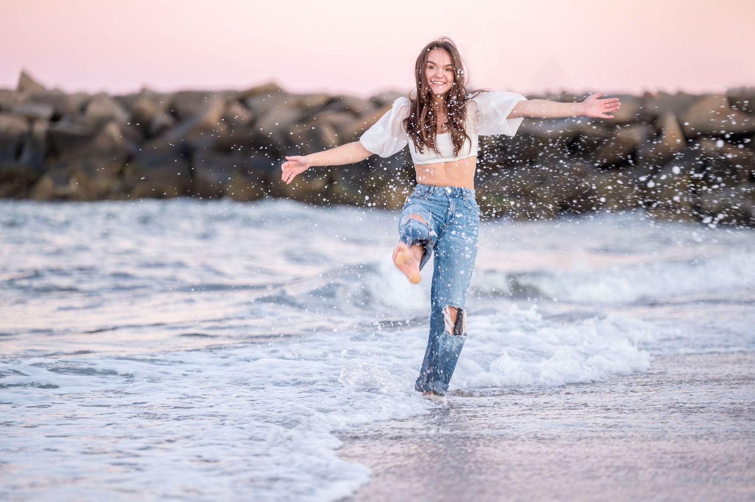 A person in white bridal attire poses gracefully on a beach with a serene ocean backdrop.