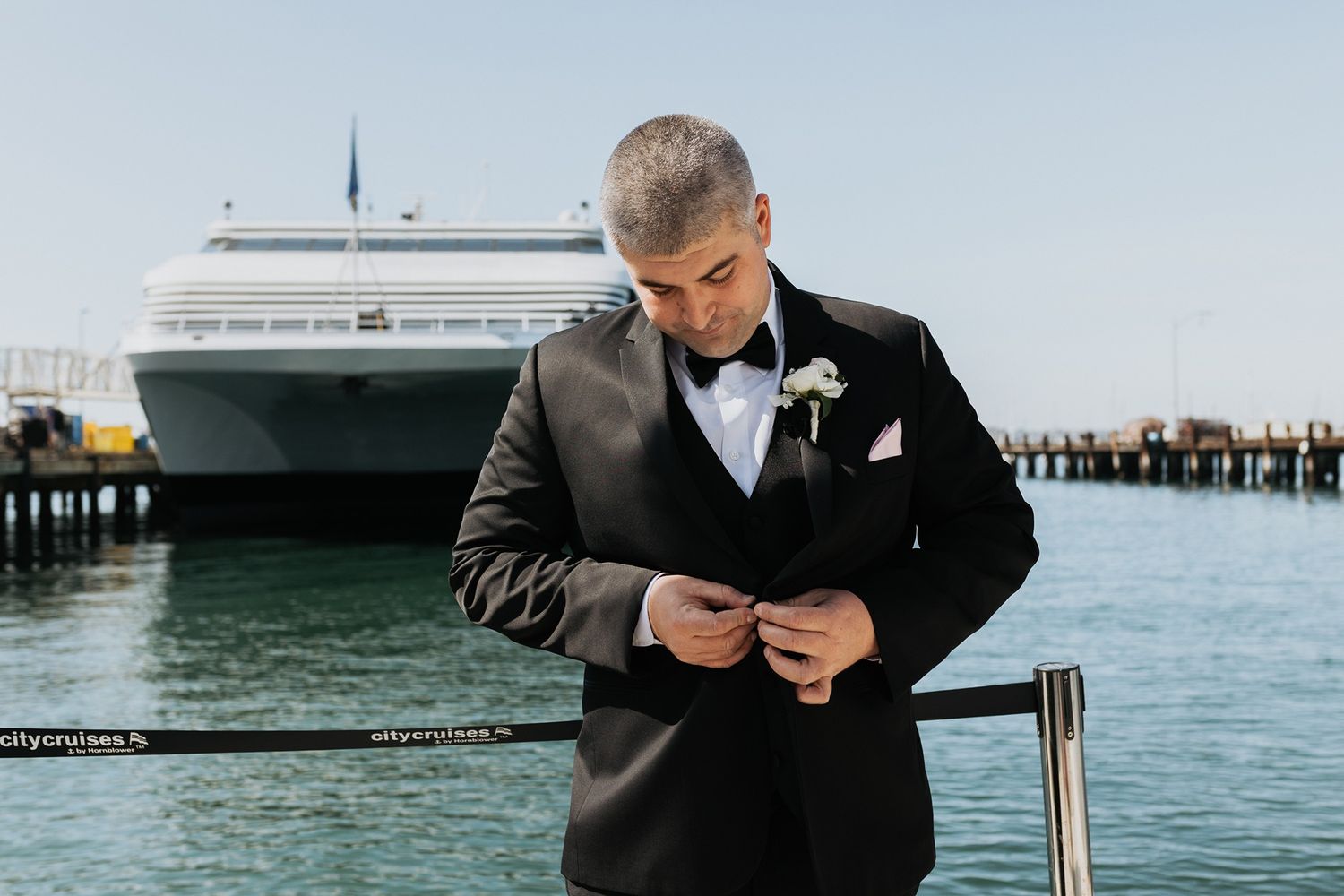 A groom in a black suits adjusts his jacket with a boat and water in the background in San Diego.