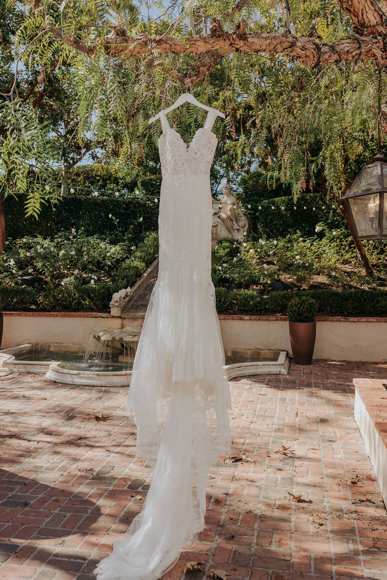 White wedding dress hanging outdoors on a sunny patio with brick pavers and lush garden greenery in the background at Rancho Bernardo Inn.