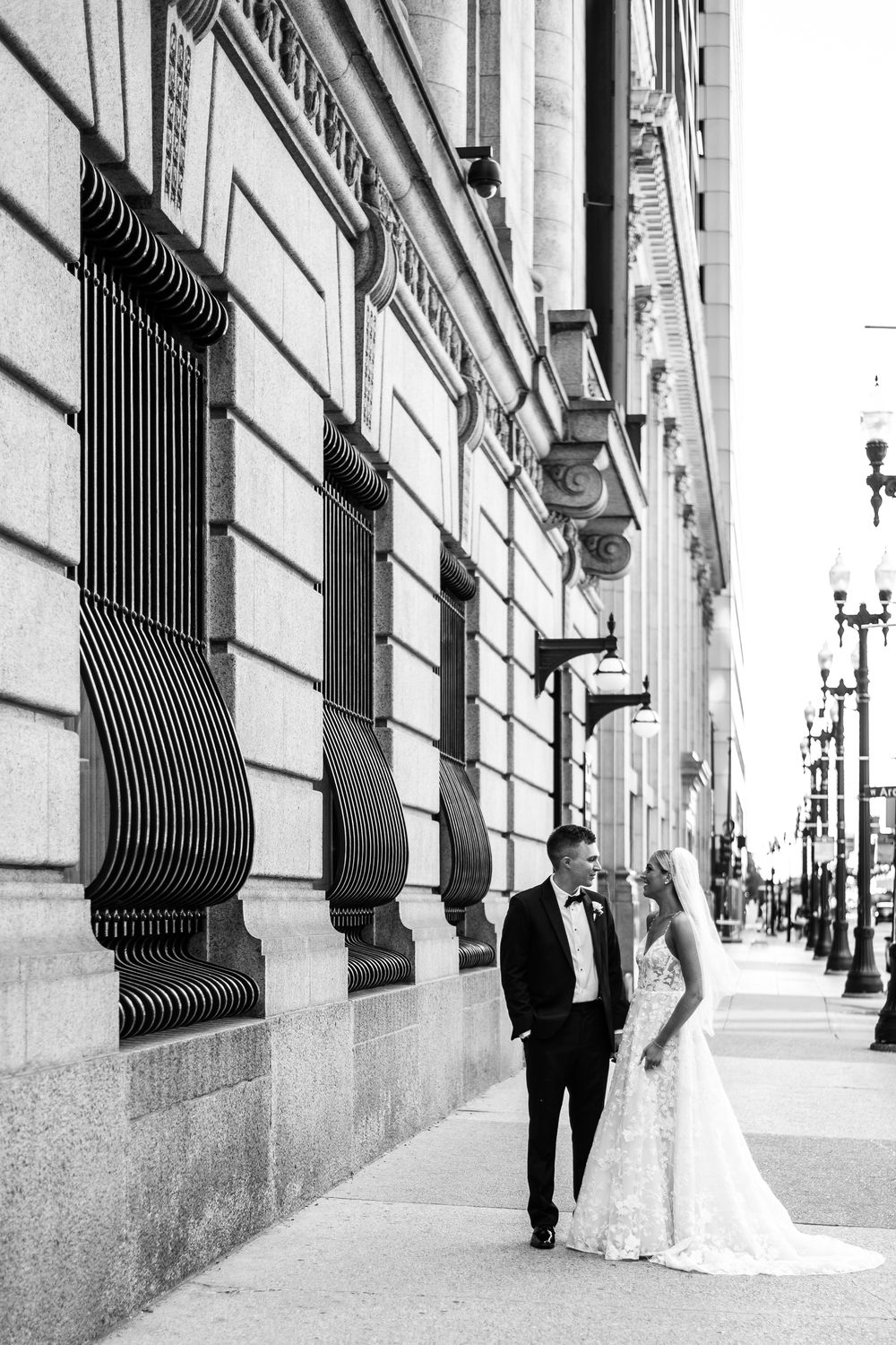 Wedding couple poses against an elegant stone building with iron window bars in an urban setting.