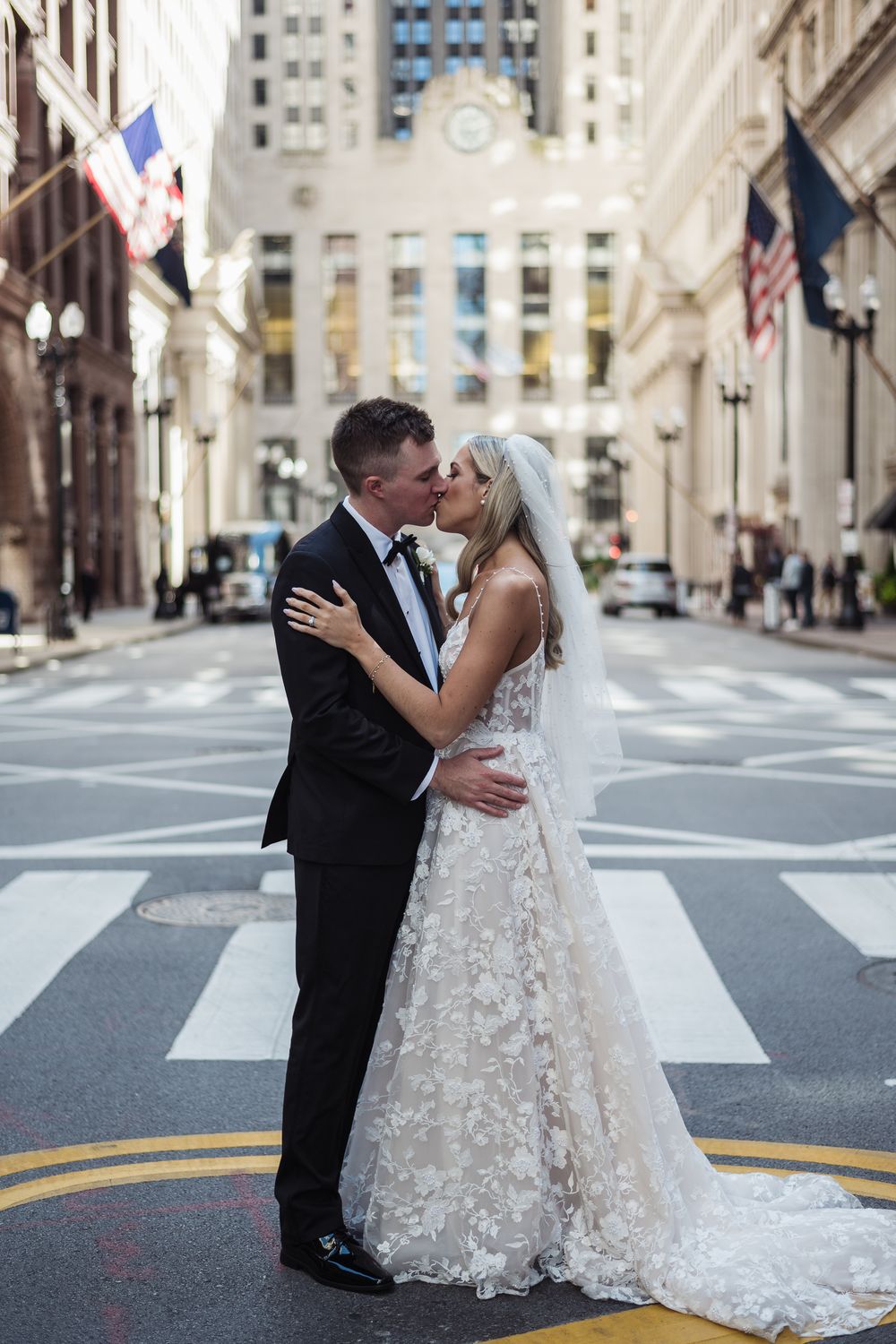 A newlywed couple shares romantic kisses on a city street with historic architecture in the background.