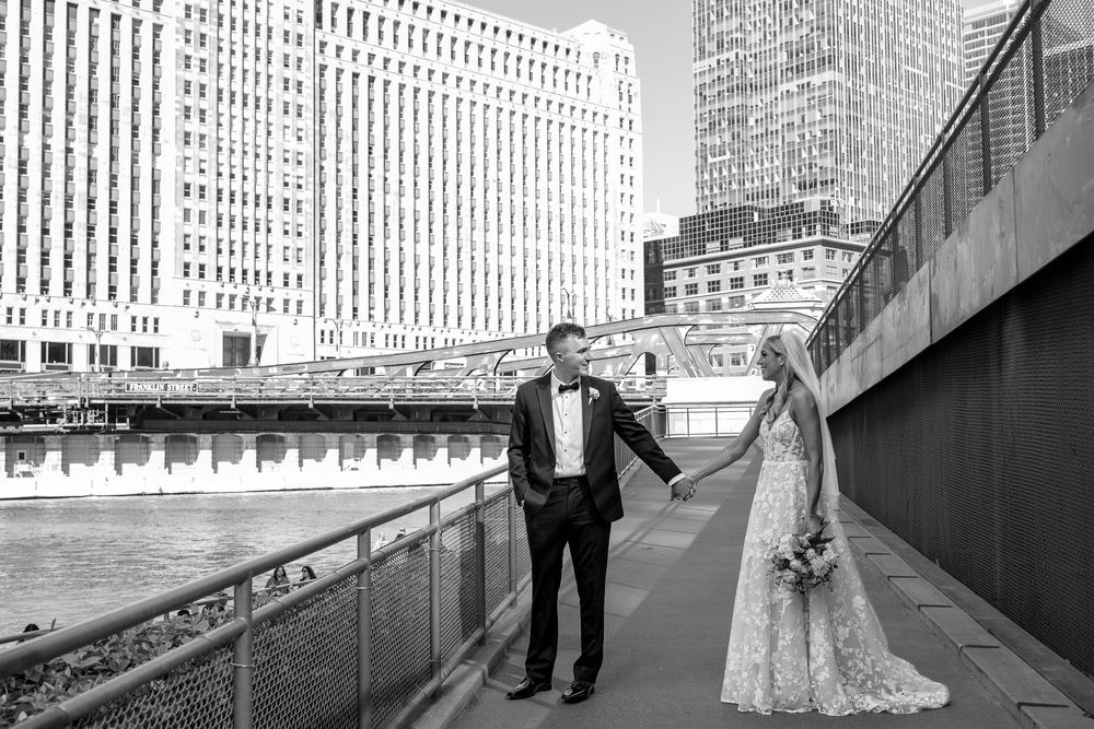 A black and white photo of a couple in wedding attire holding hands along an urban riverside walkway in Chicago.