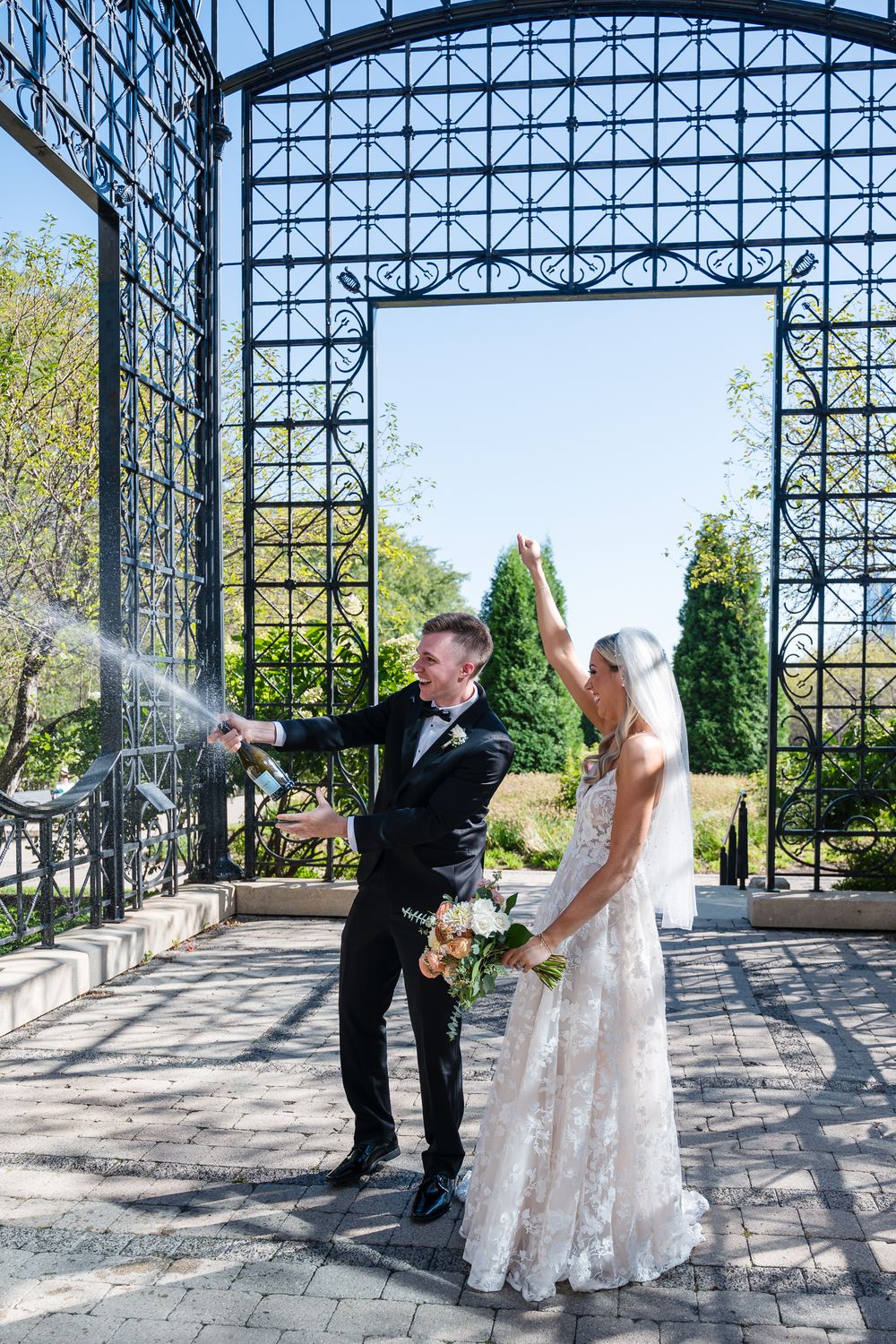 A couple shares a playful moment during their wedding photoshoot under an ornate steel and glass pavilion structure.