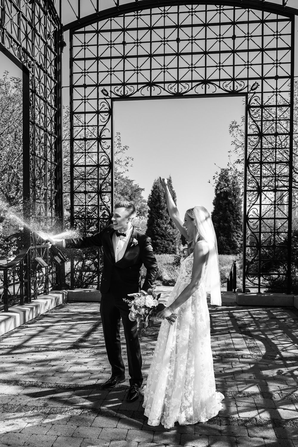 A black and white wedding photo showing a couple standing under an ornate metal archway in a garden setting.