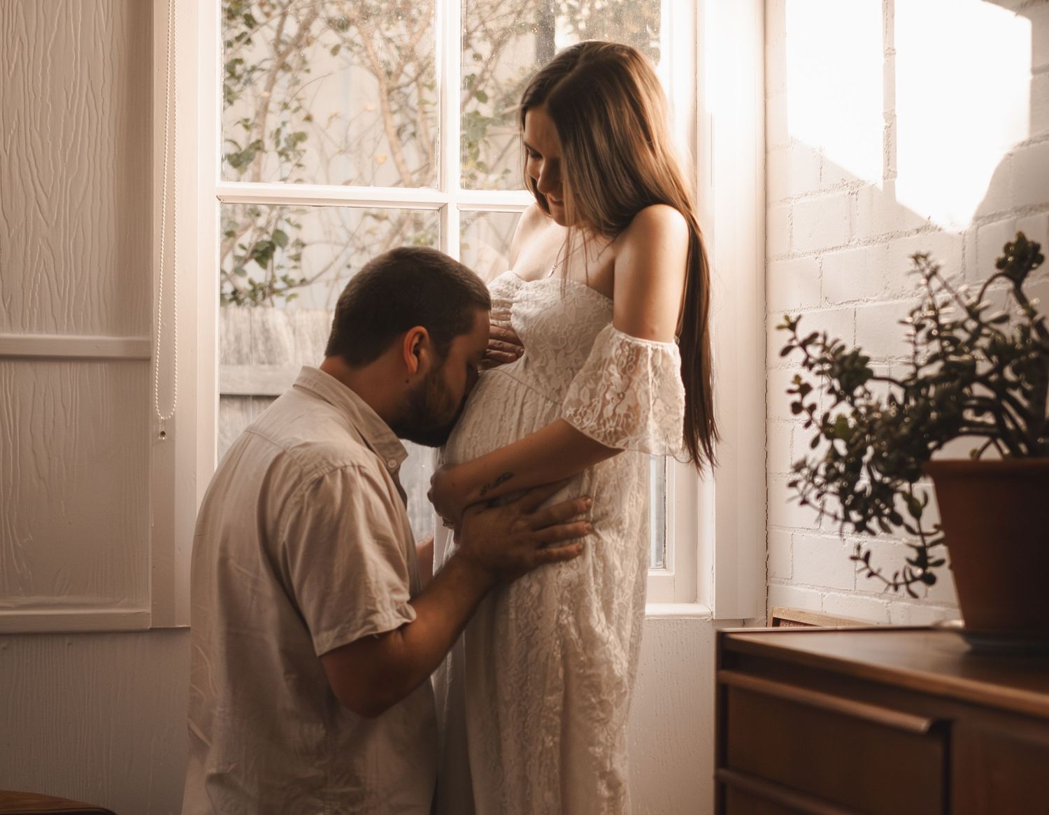 A man kneels before a pregnant woman in a white lace dress by a sunlit window indoors.
