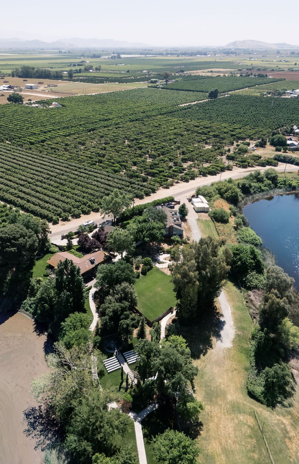 Aerial view of rustic wedding venue surrounded by vineyards, orchards, and a serene pond in rural countryside.