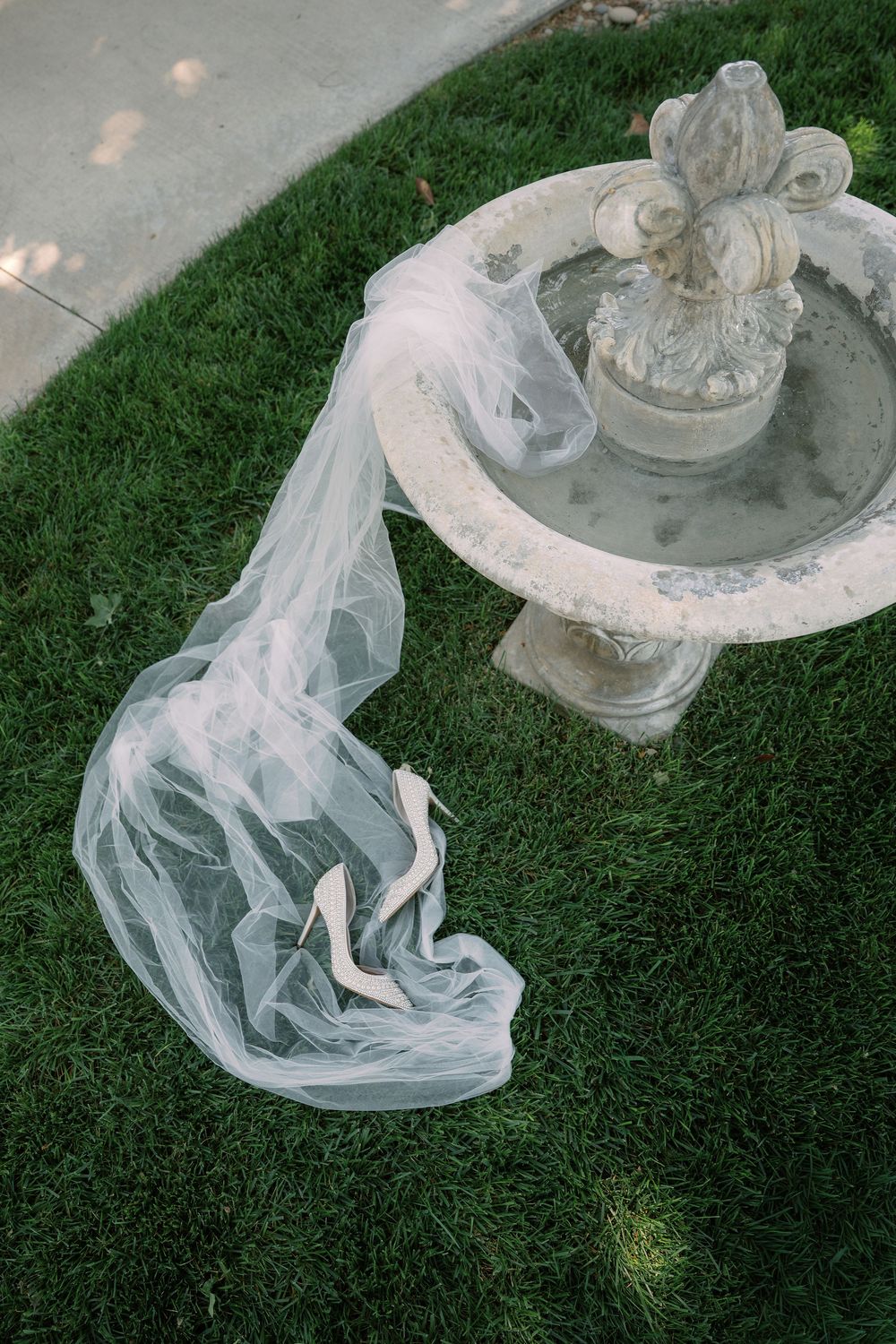 Bridal veil and white heels draped over a stone garden fountain on lush green grass.