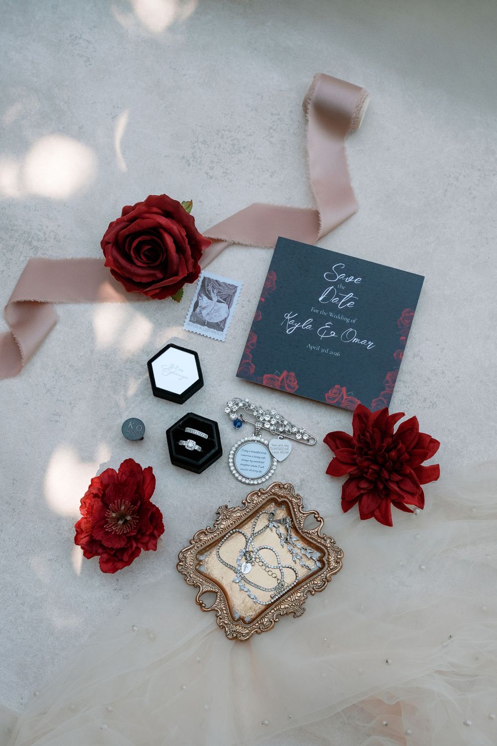 Elegant wedding flat lay with save the date card, diamond ring, red roses, pearl jewelry, and blush ribbon on white surface.
