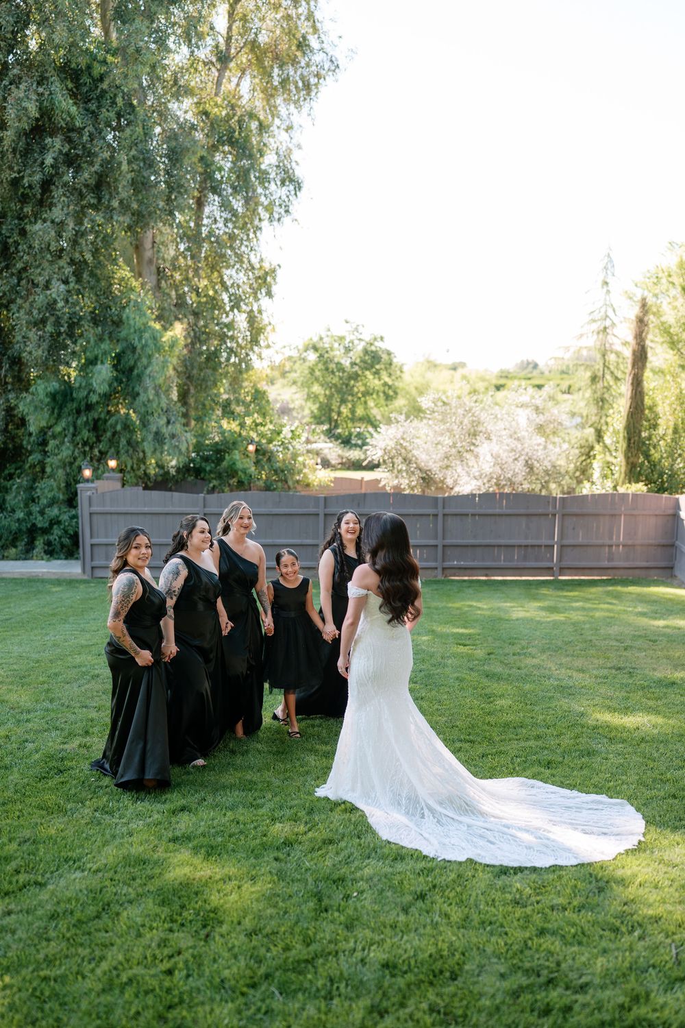 Bride in white gown reveals dress to bridesmaids in black dresses on green lawn.