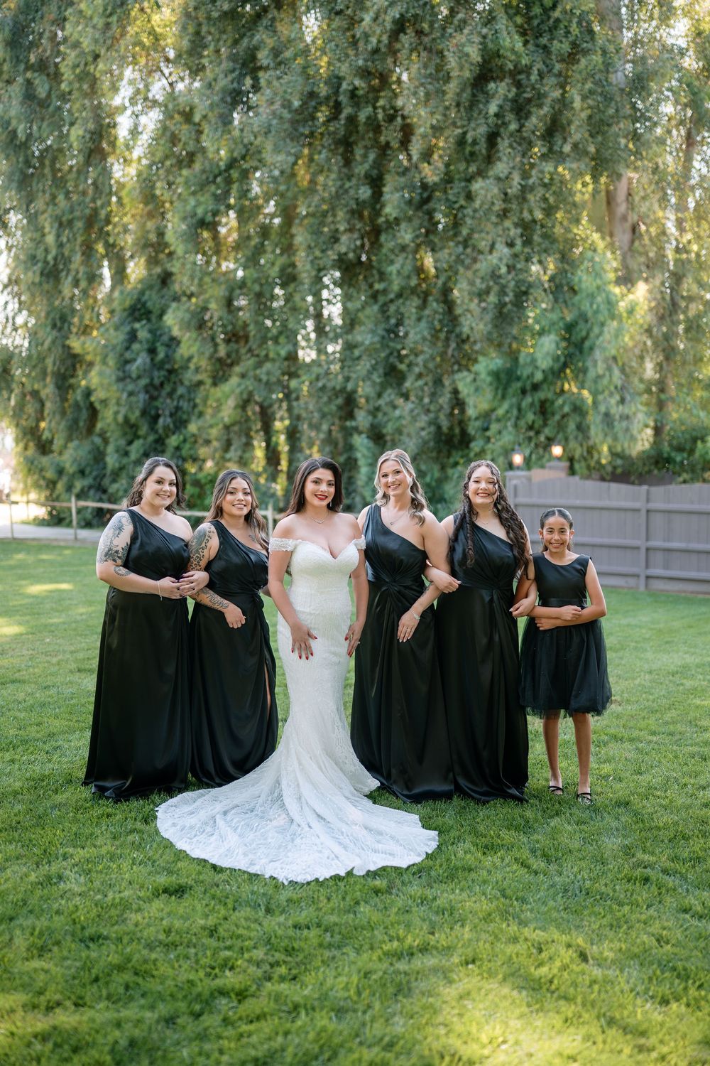 Bride in white gown poses with bridesmaids in black dresses on green lawn before weeping willow tree.