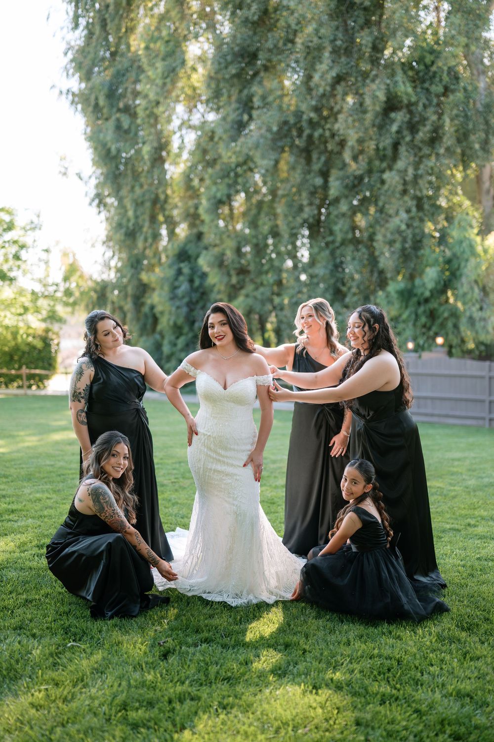 Bride in white lace gown surrounded by bridesmaids in black dresses on green lawn near weeping willow.