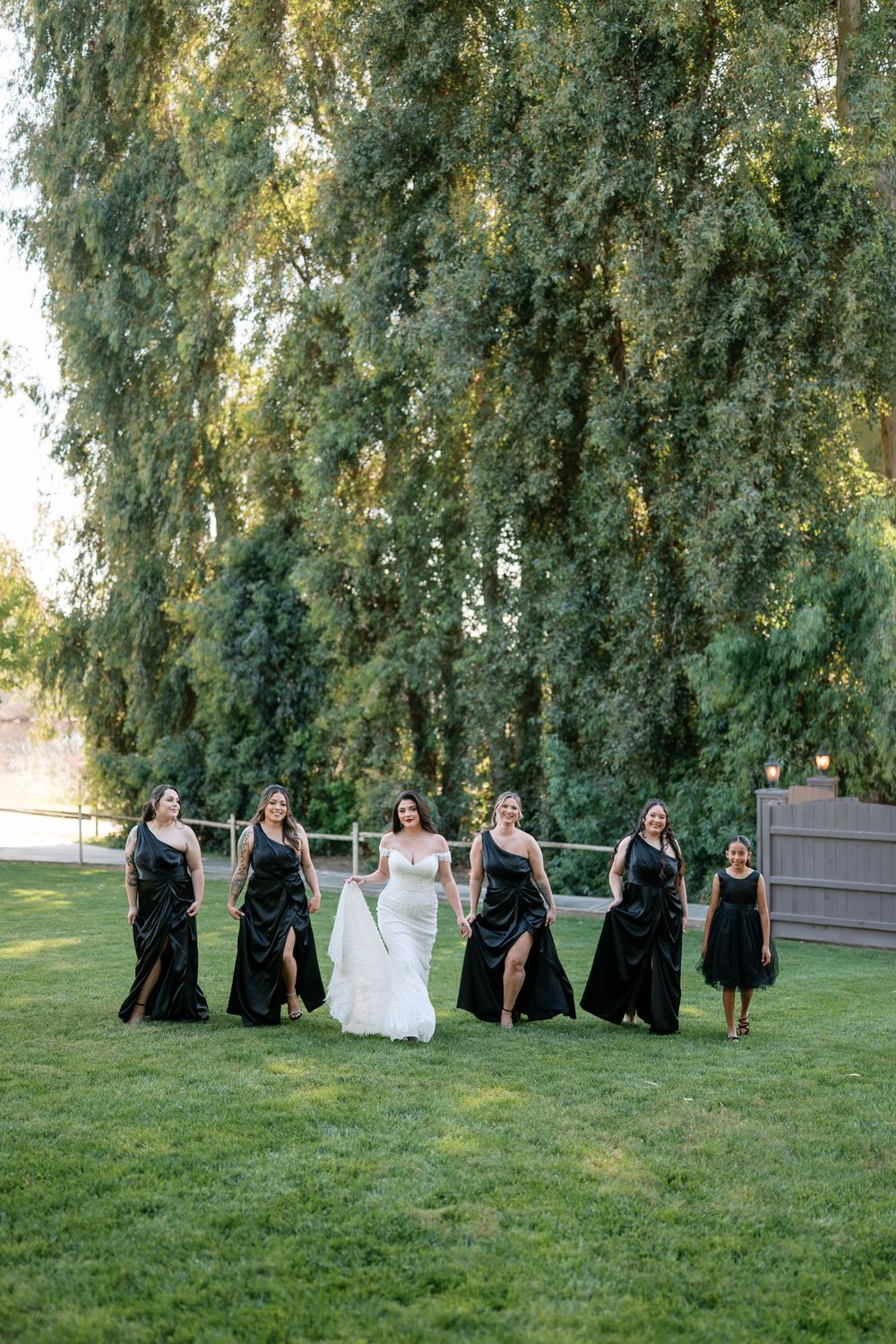 Bride in white gown walking with bridesmaids in elegant black dresses across lush green lawn at sunset.