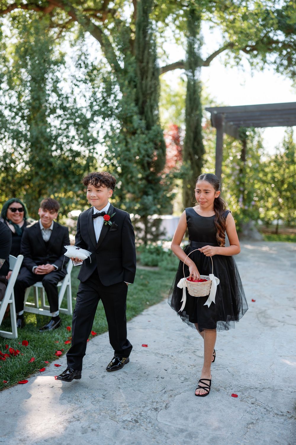 Ring bearer and flower girl in black attire walk down outdoor wedding aisle, scattering red rose petals.
