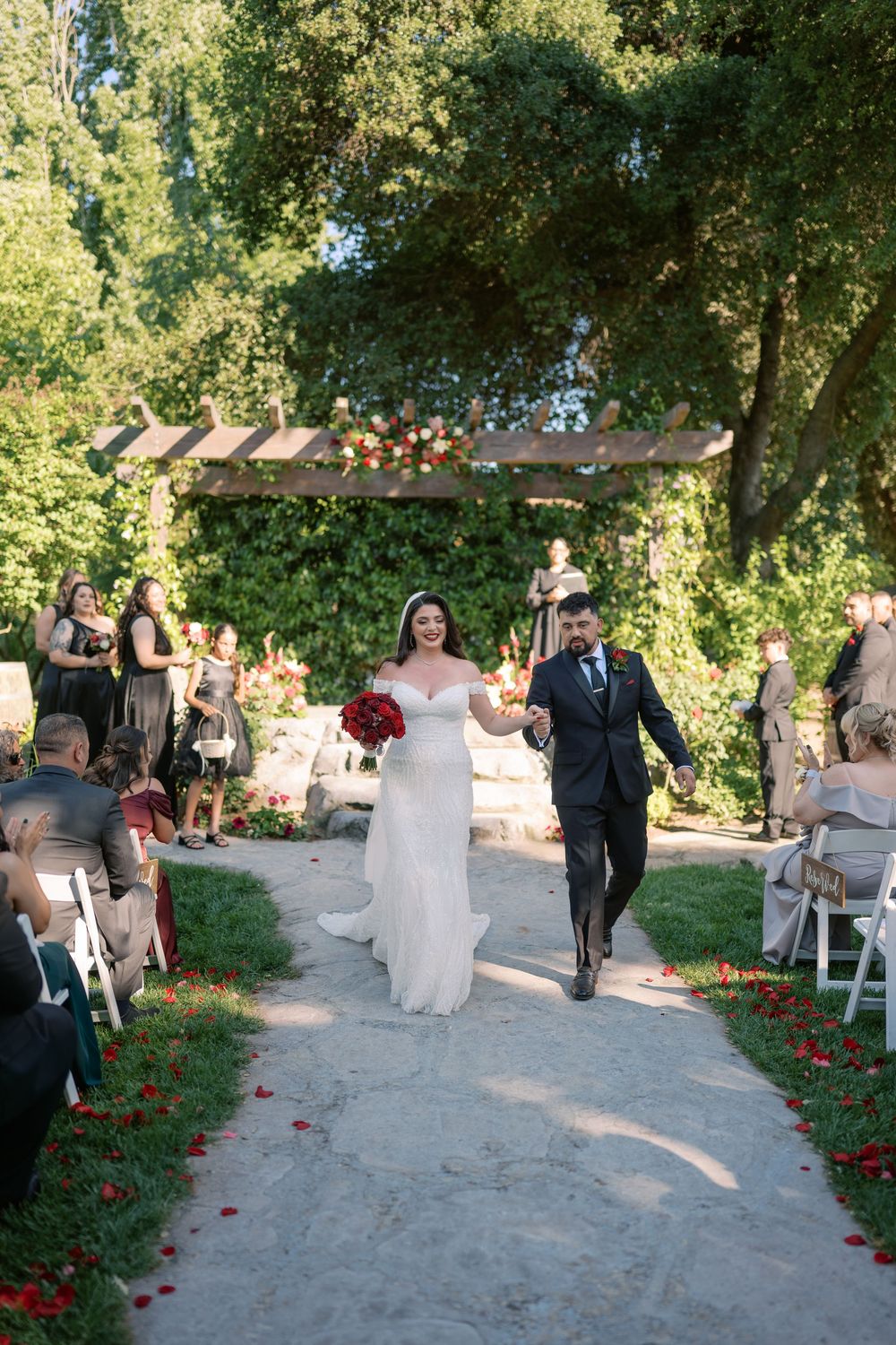 Newlywed couple walks joyfully down outdoor aisle after wedding ceremony, bride holding red bouquet.