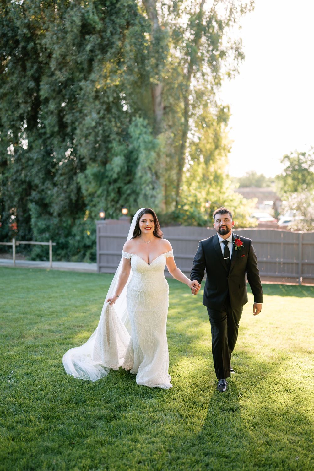 Bride in off-shoulder gown and groom in black suit hold hands walking on green lawn at golden hour.