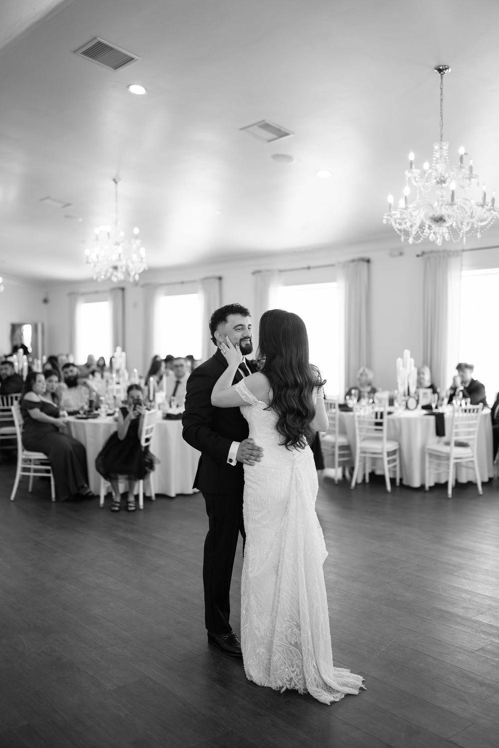 Bride and groom share first dance in elegant ballroom with crystal chandeliers and seated wedding guests.