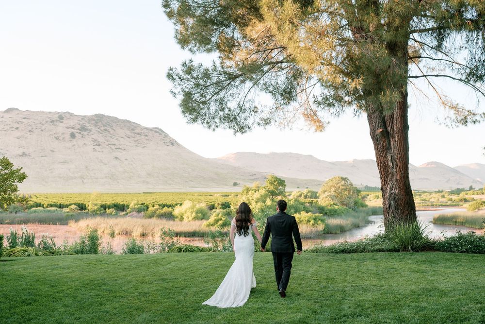 Bride and groom walk hand-in-hand across lush green lawn toward scenic mountain and river landscape at golden hour.