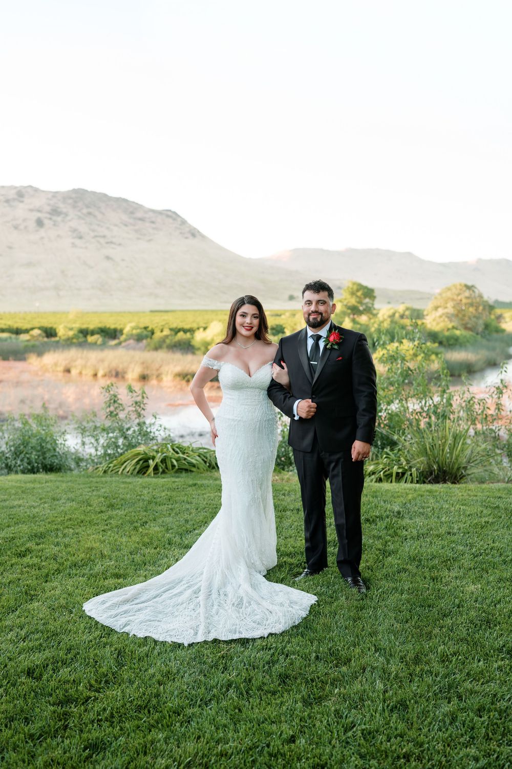 Bride in fitted lace gown and groom in black suit pose together on grass with mountain backdrop.