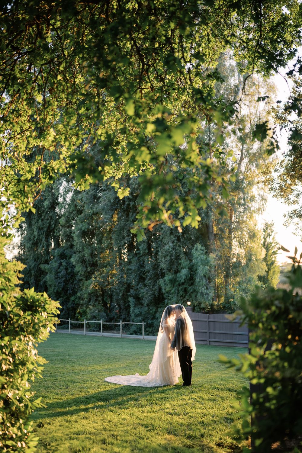 Bride and groom share intimate moment framed by lush green trees on manicured lawn at sunset.