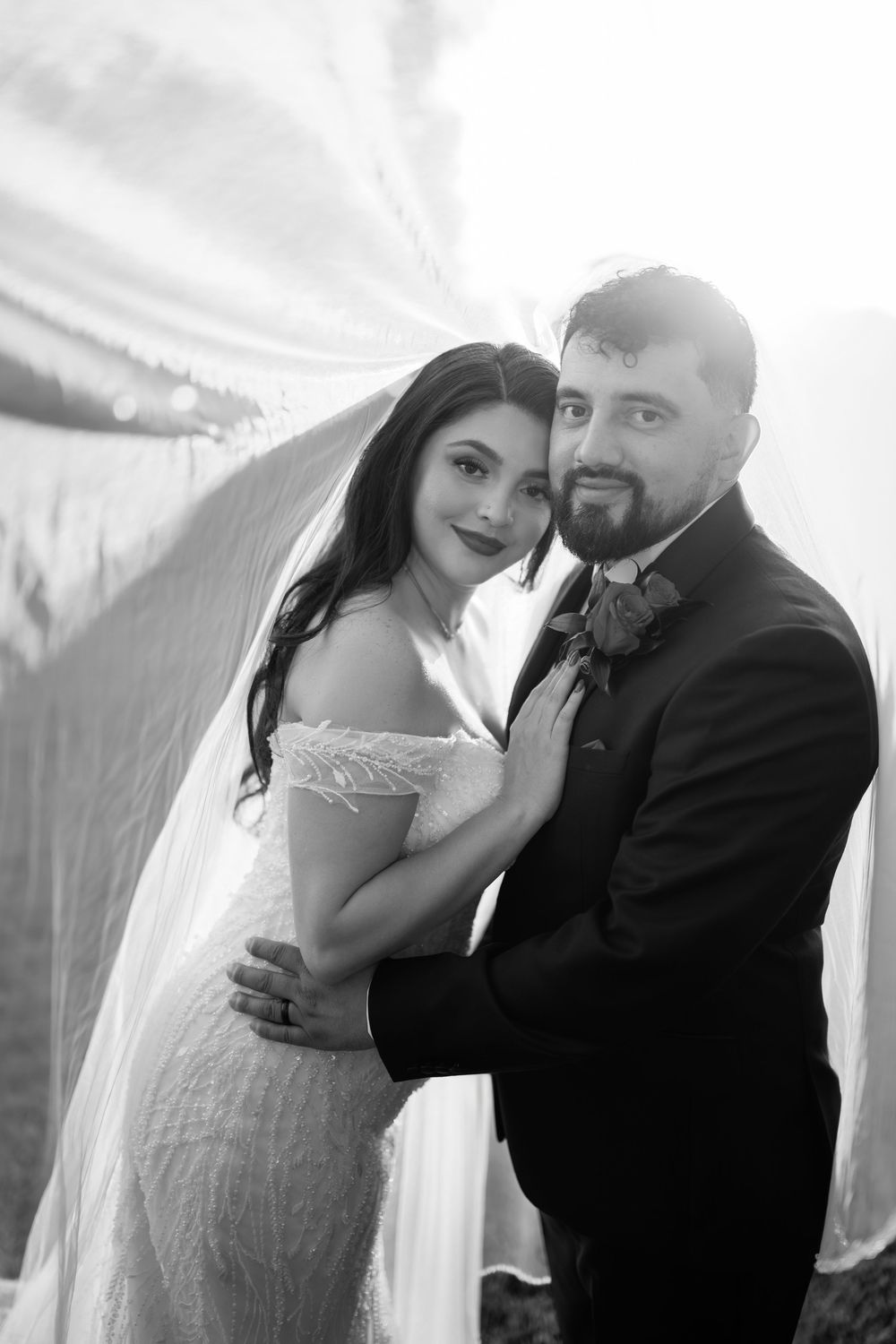 Black and white portrait of bride and groom embracing under flowing veil on their wedding day.