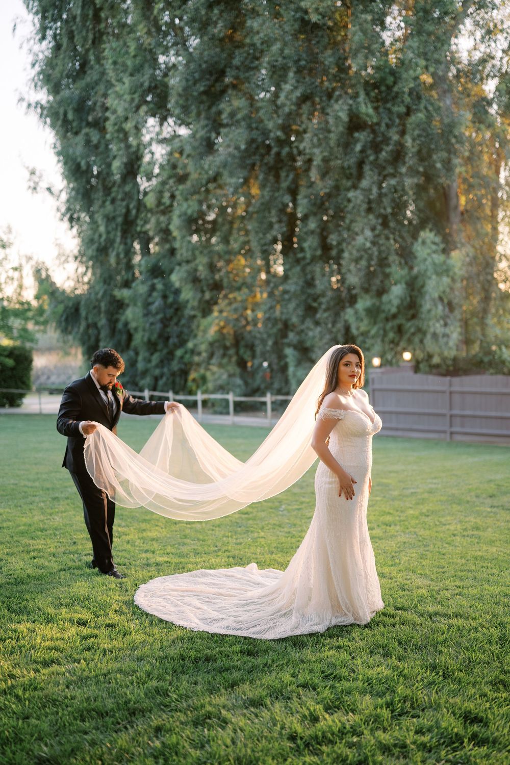 Bride in white mermaid gown with long veil held by groom in black suit on green lawn with trees.