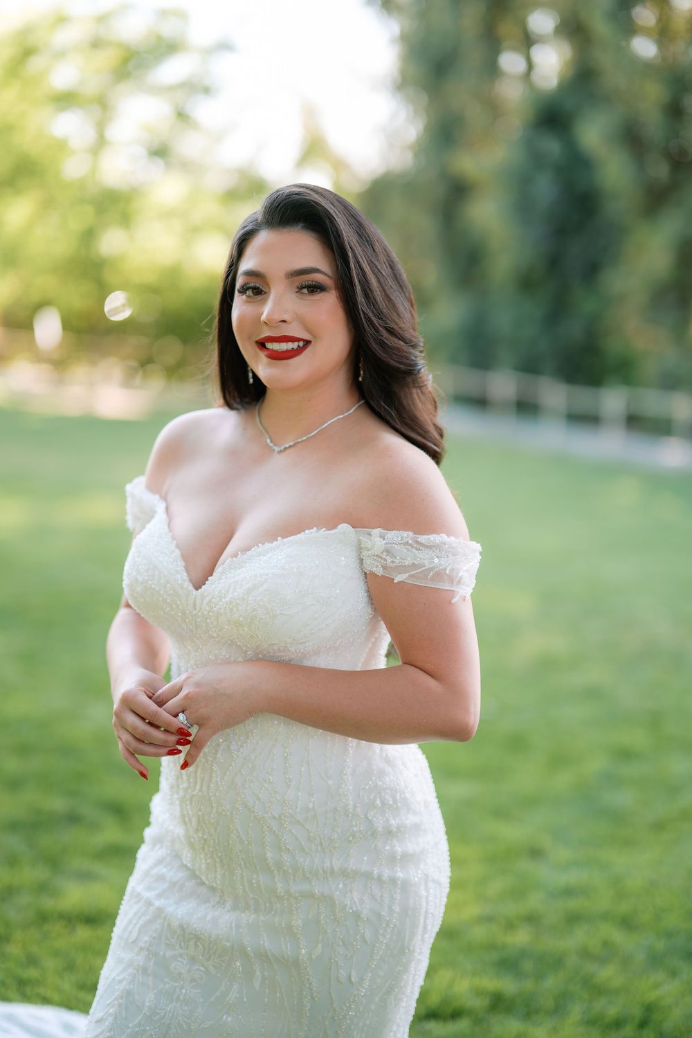 Smiling bride in off-shoulder white lace wedding dress with red lips and necklace outdoors.
