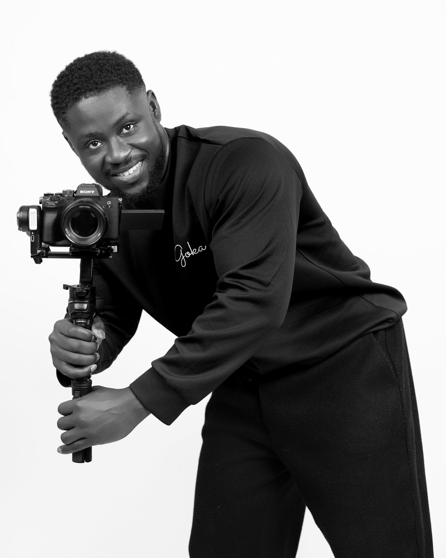 Black and white photo of a smiling man in black outfit holding a camera on a gimbal stabilizer.