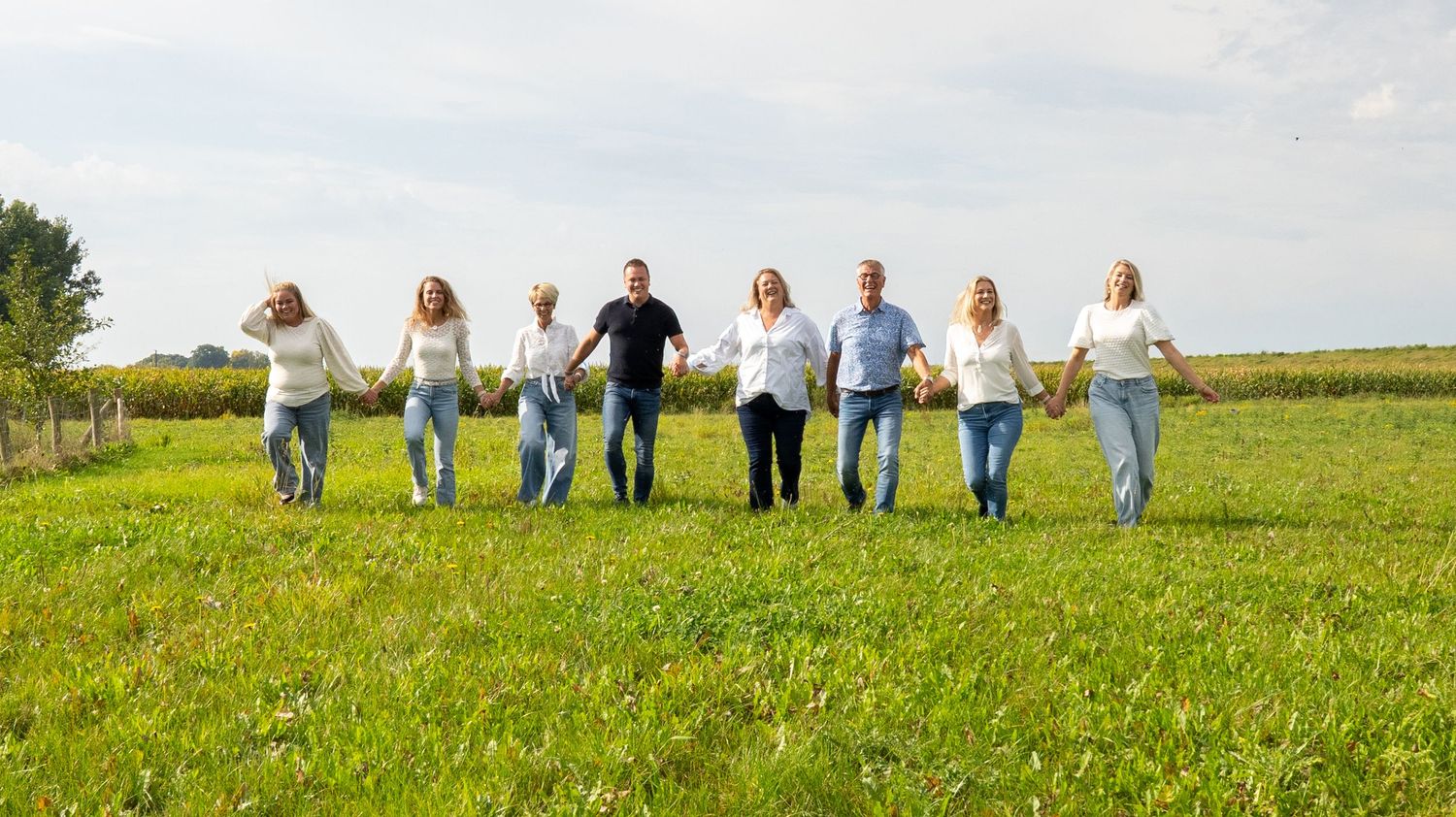 Een groep van acht mensen loopt hand in hand door een groene weide onder een bewolkte hemel, gekleed in casual witte kleding. familiefoto