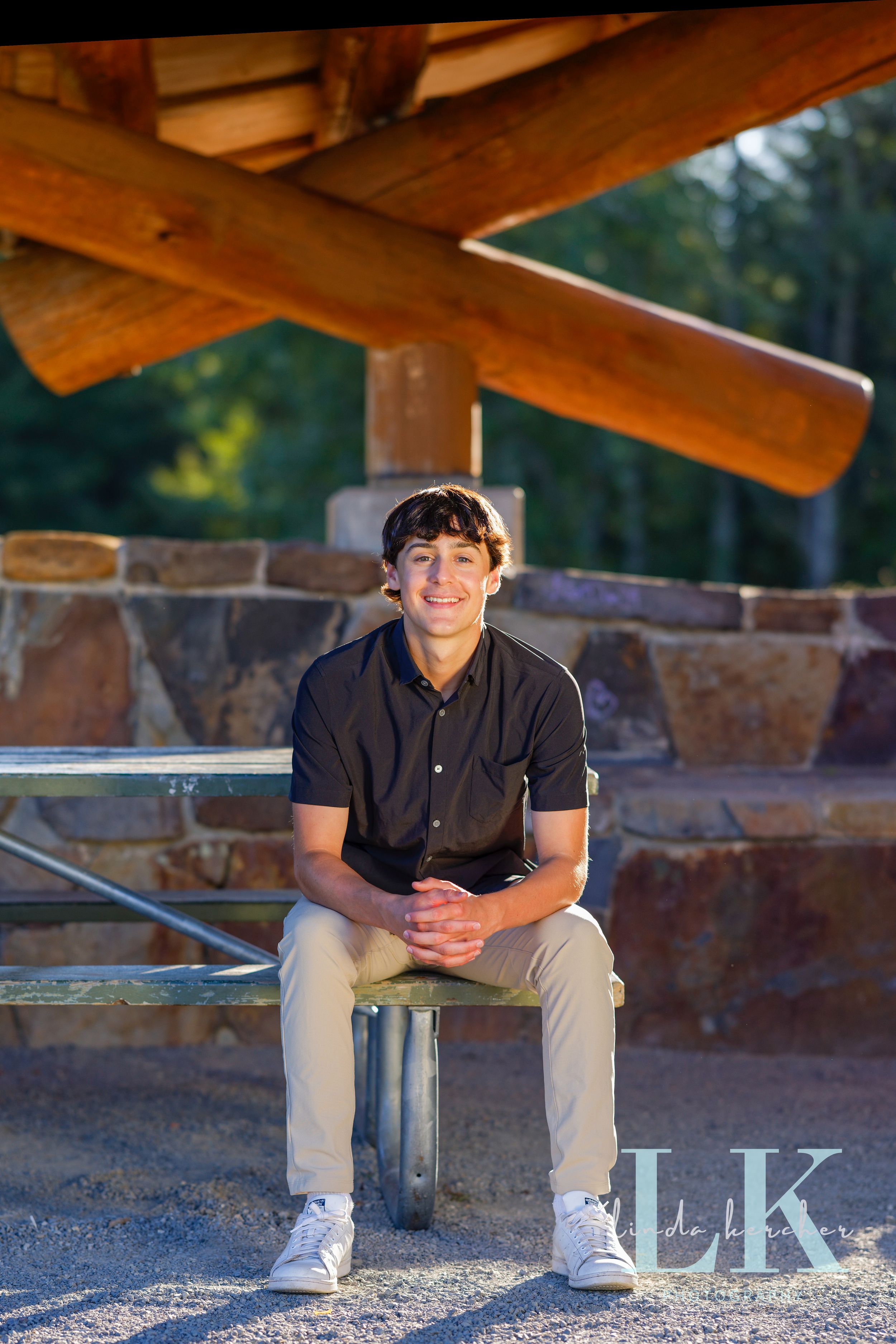 Teen boy in dark polo and khakis sits smiling under a rustic wooden pavilion for senior portraits.