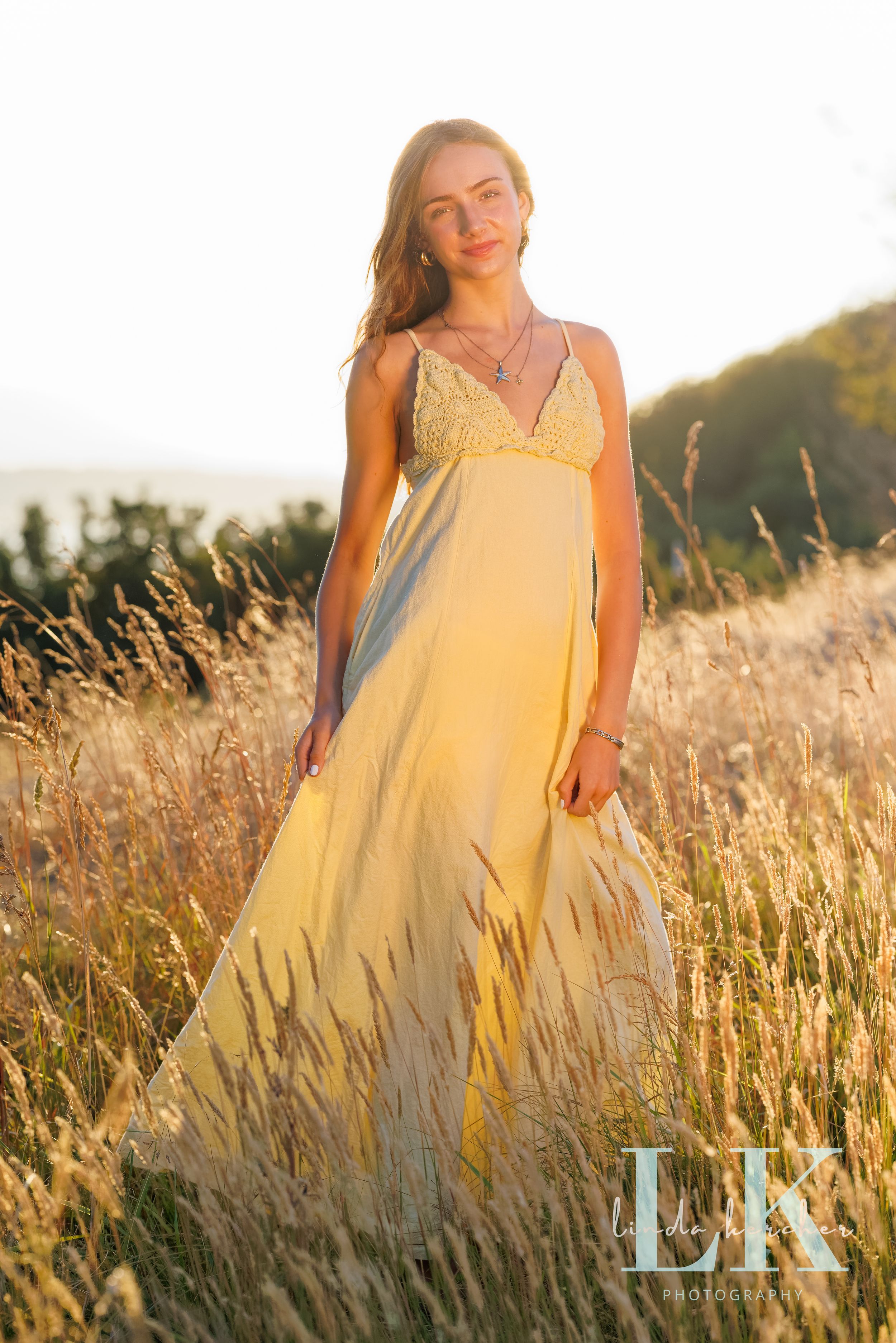 Young woman in yellow maxi dress standing in golden meadow grass during sunset, professional senior portrait photography.