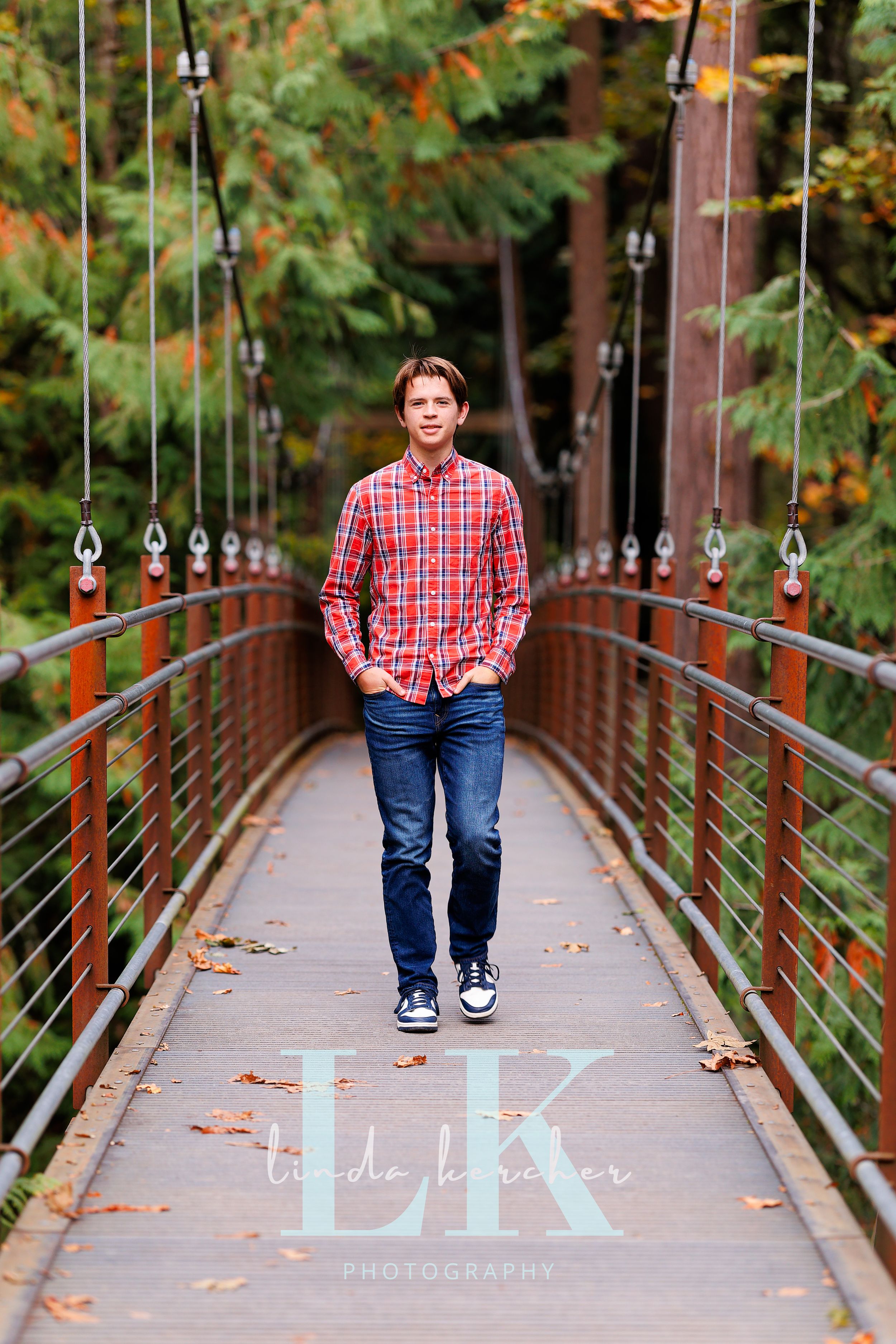 Teen boy in red plaid shirt and jeans stands on a suspension bridge surrounded by fall foliage.