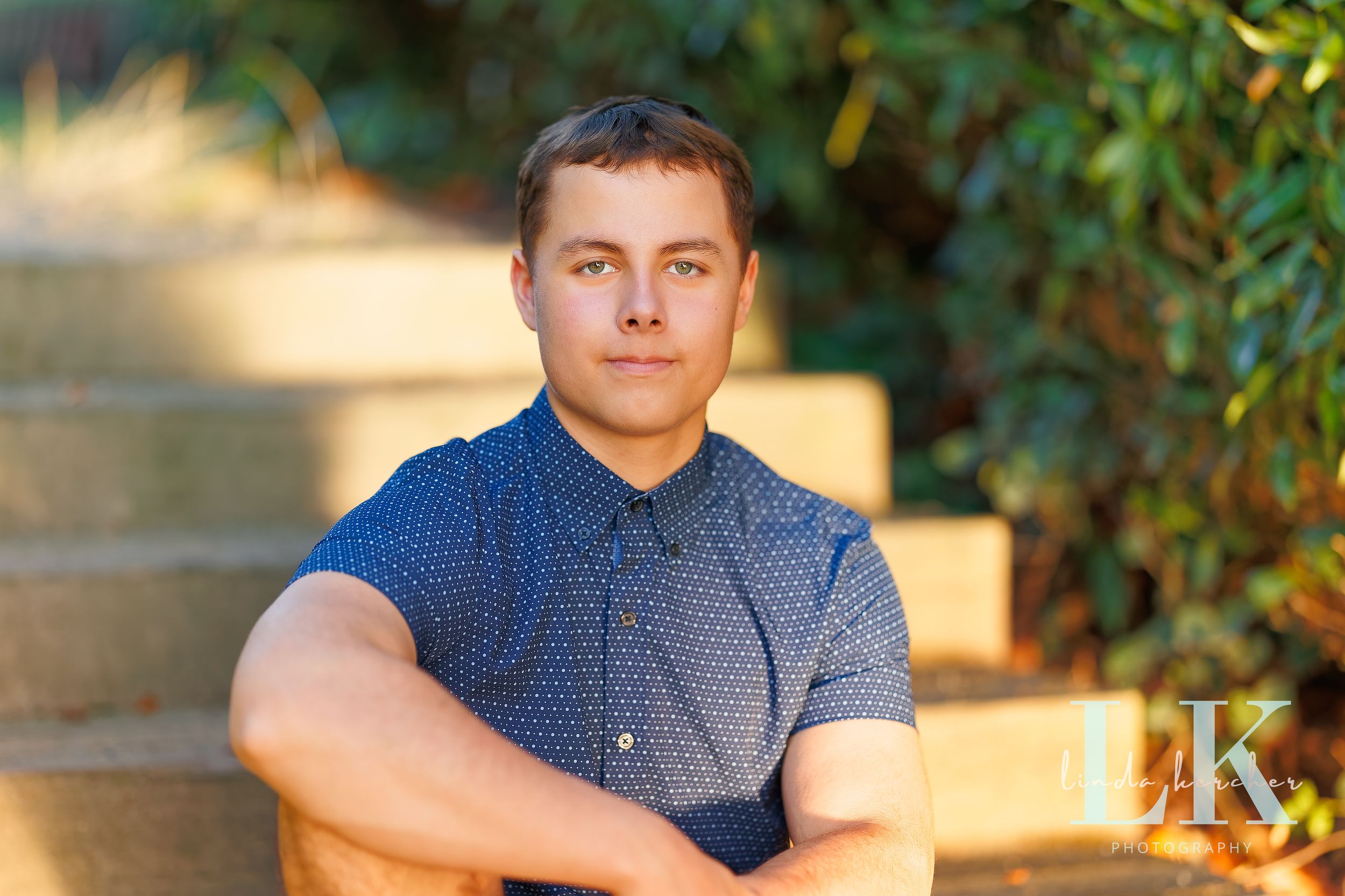 Young man in blue patterned shirt sitting with arms crossed outdoors on stone steps surrounded by greenery.