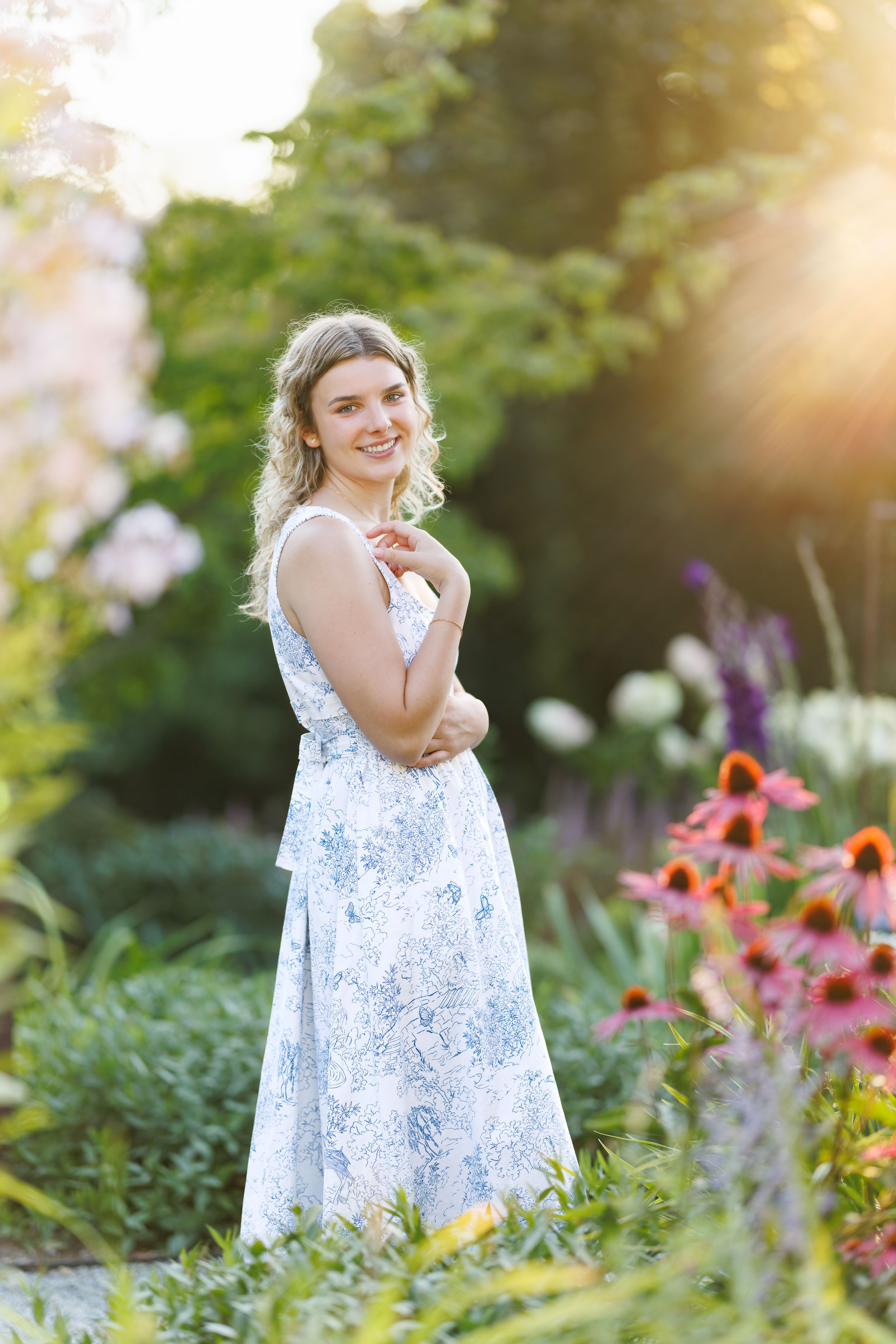 Smiling blonde woman in floral maxi dress posing in a colorful sunlit flower garden.