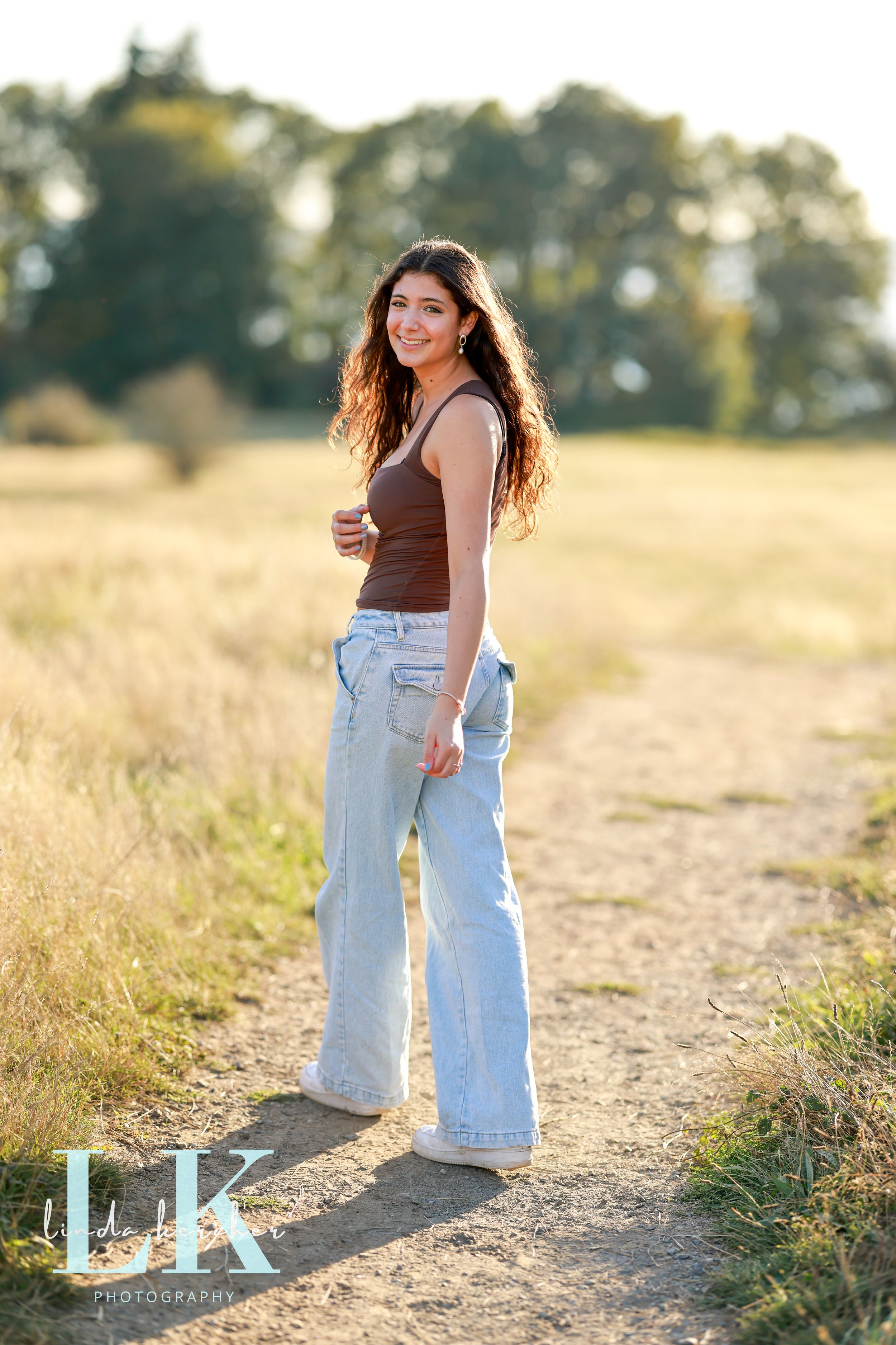 Smiling brunette woman in brown tank top and wide-leg jeans on a sunny dirt path.
