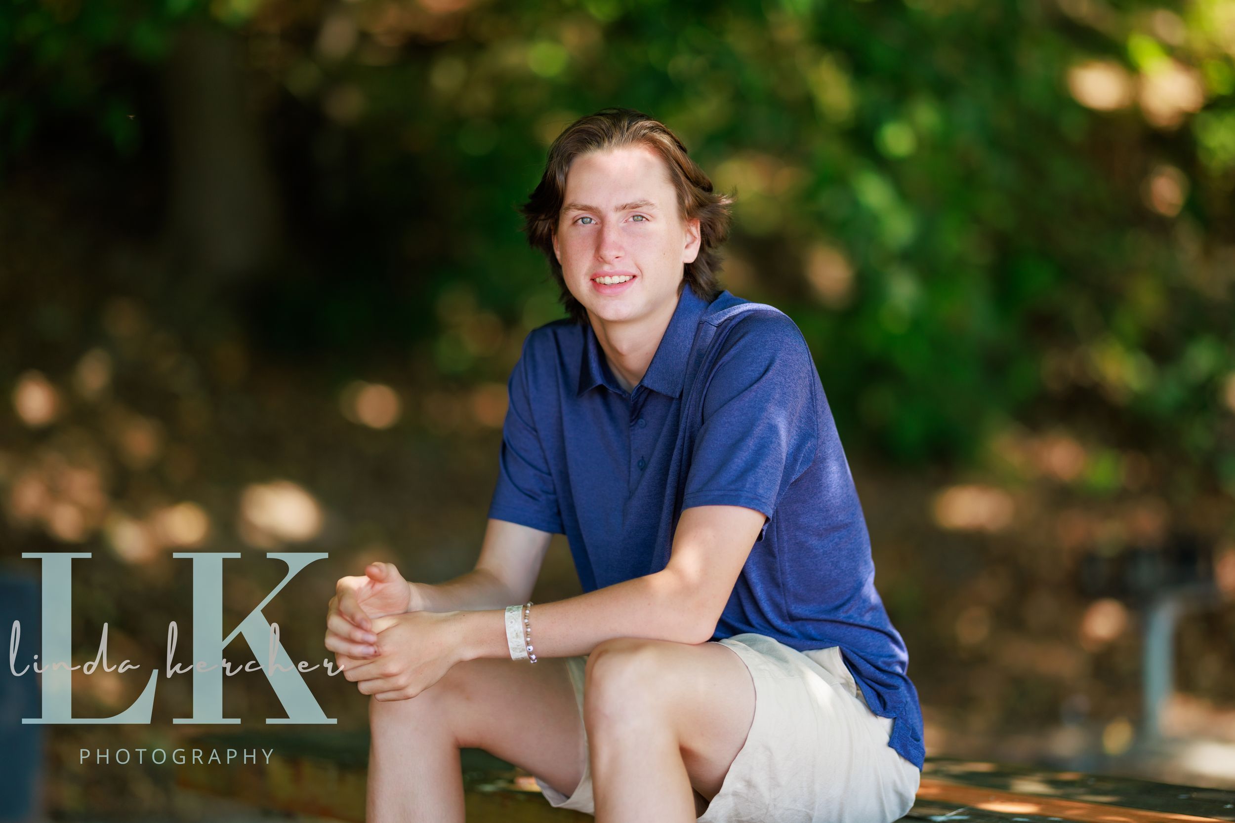 Young man in blue polo shirt and white shorts smiling outdoors in a natural wooded setting, senior portrait photo.
