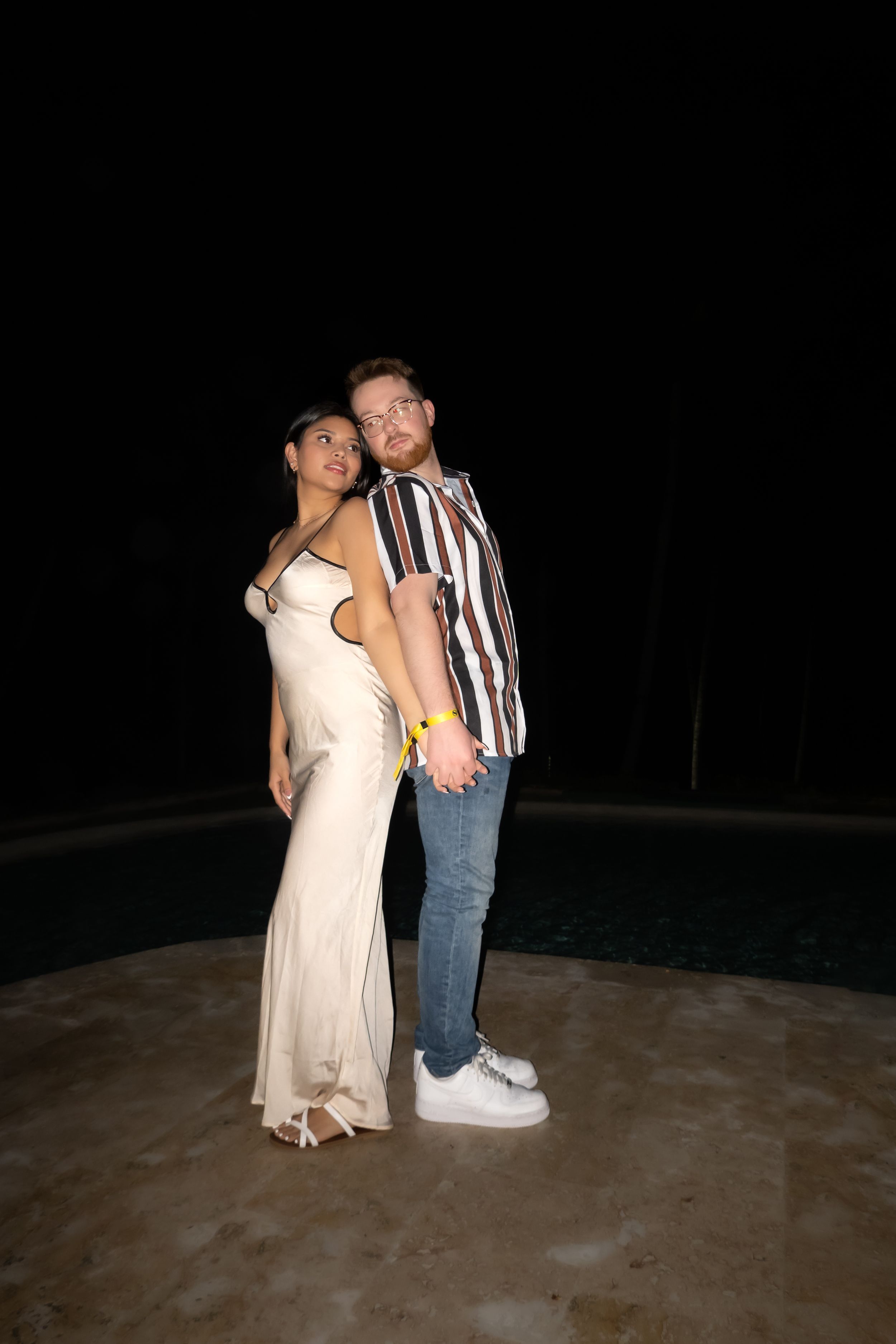 Portrait of someone in a striped short-sleeve button down shirt and khaki pants standing by a pool at dusk.