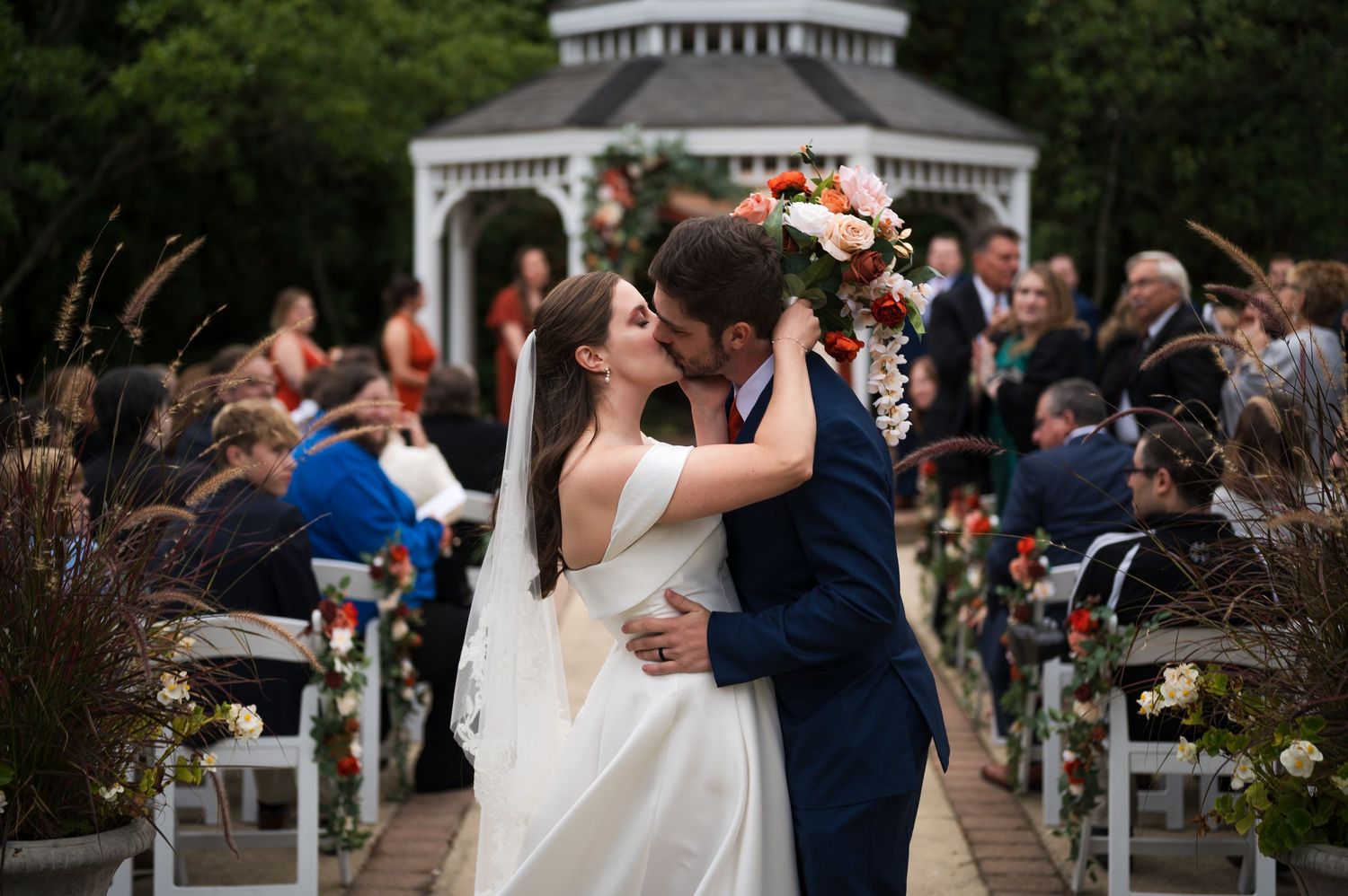 A romantic wedding ceremony takes place at an outdoor garden venue with a white gazebo and decorated aisle.