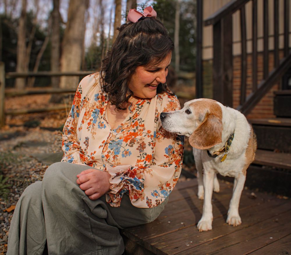 A person in a floral dress sits on wooden steps with a white and brown dog on a porch.