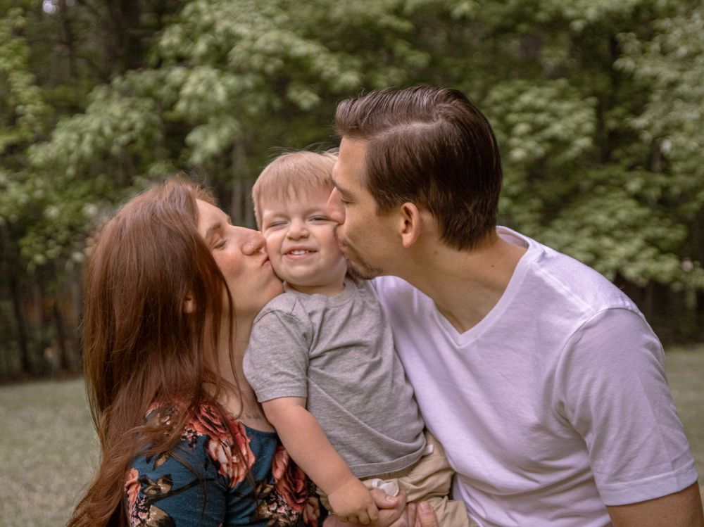 Parents kissing their smiling toddler boy on cheeks outdoors in a lush green park setting.