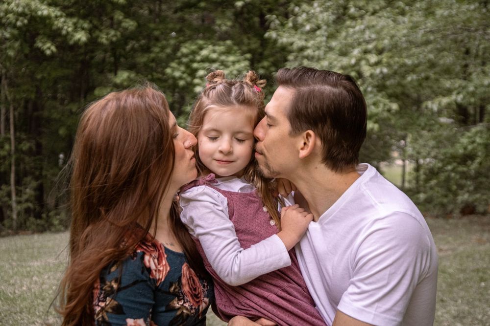 Parents kissing their young daughter on cheeks outdoors in a green park during a family photo session.