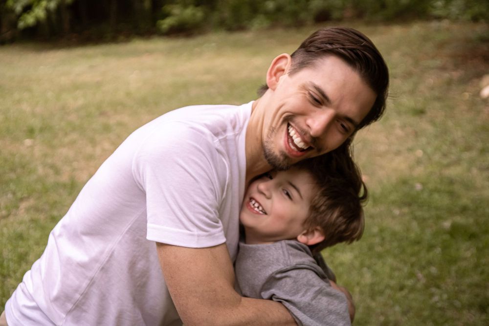 A laughing father hugs his smiling young son outdoors on a green grass lawn in a heartwarming candid family moment.