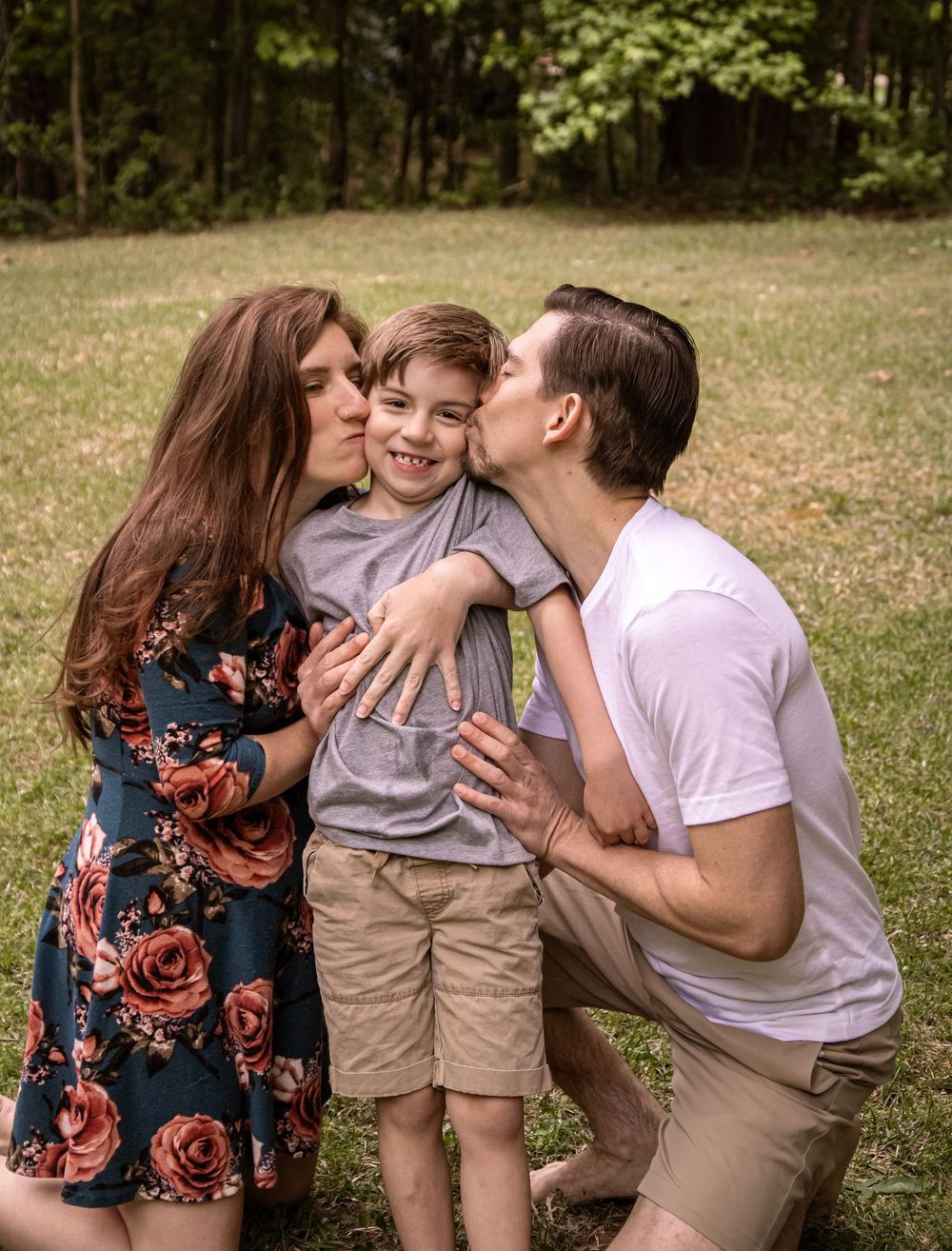 Parents kissing their young son on cheeks while posing together on green grass during outdoor family photos.